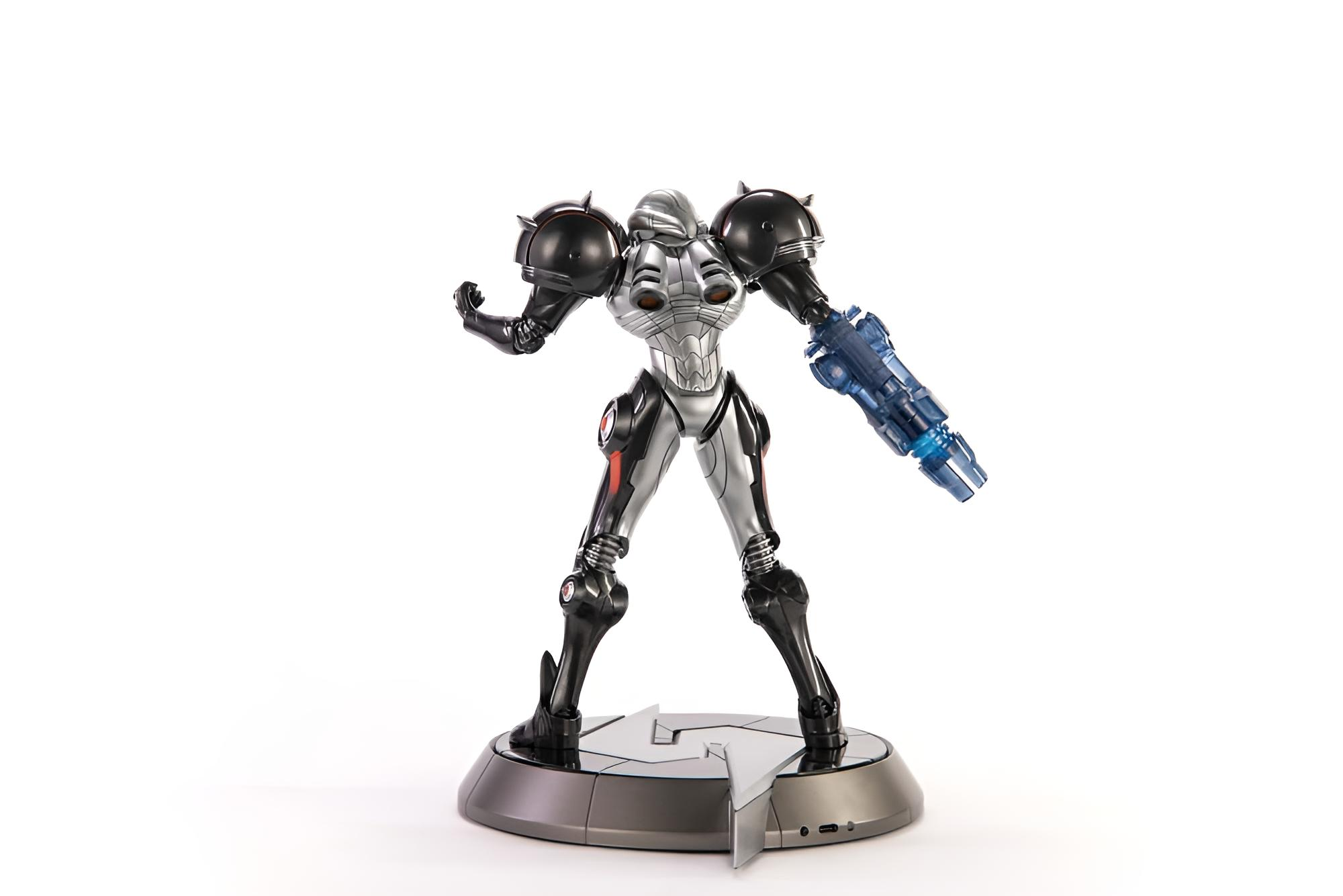 Eine silber-schwarze Actionfigur mit blauer Armkanone steht auf einem runden Sockel vor weißem Hintergrund.