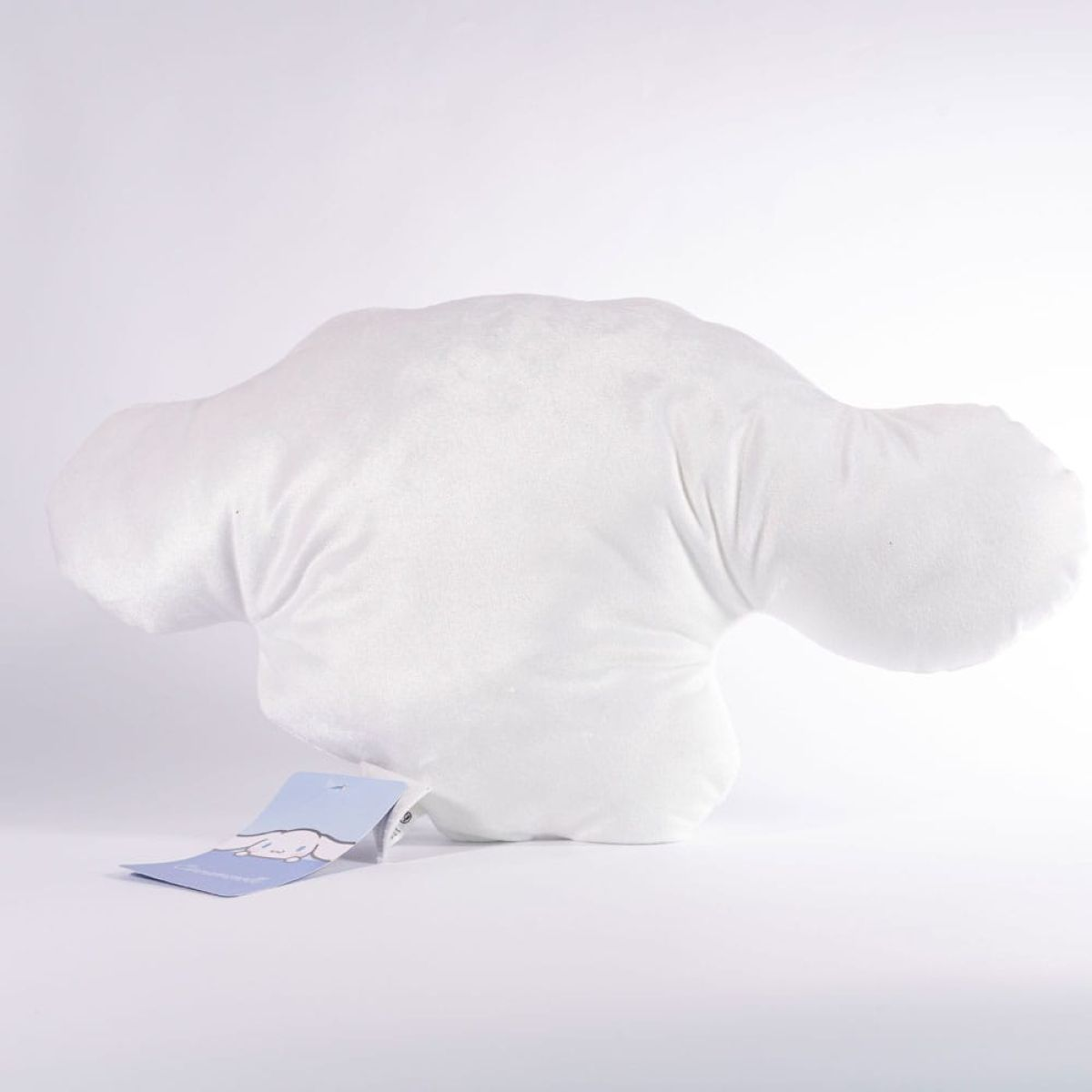 Un cojín de felpa blanco con forma de cabeza de personaje y orejas largas sobre superficie blanca con etiqueta.