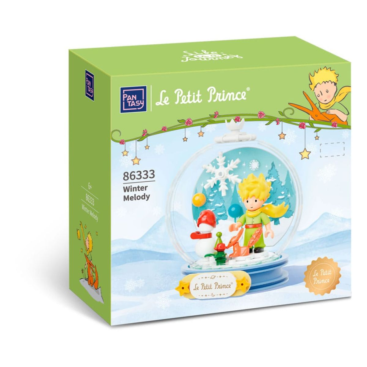 Eine grüne Spielzeugbox zeigt eine Schneekugel mit der Figur Der Kleine Prinz, einem Schneemann und Bäumen.