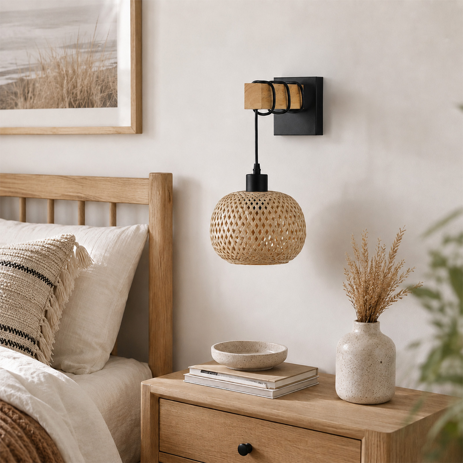 Una mesita de noche con libros, un cuenco y un florero al lado de una cama con una lámpara de pared.