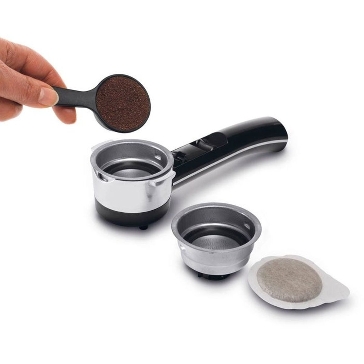 Hand füllt Kaffeepulver aus Löffel in einen Siebträger; ein weiterer Filter und Kaffeepad liegen daneben auf weißem Grund.
