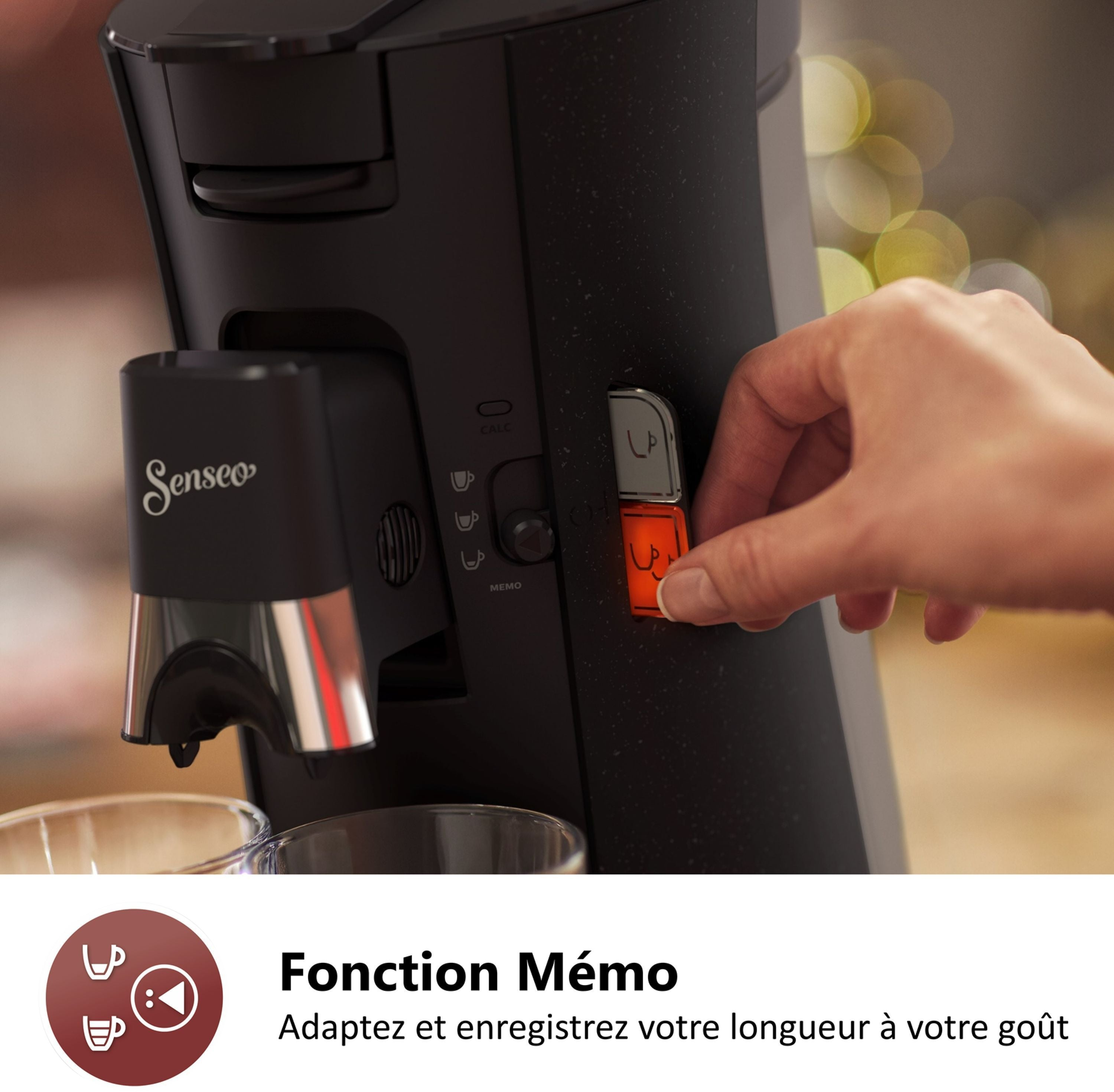 Una mano presiona el botón naranja en una cafetera Senseo negra con dos vasos.