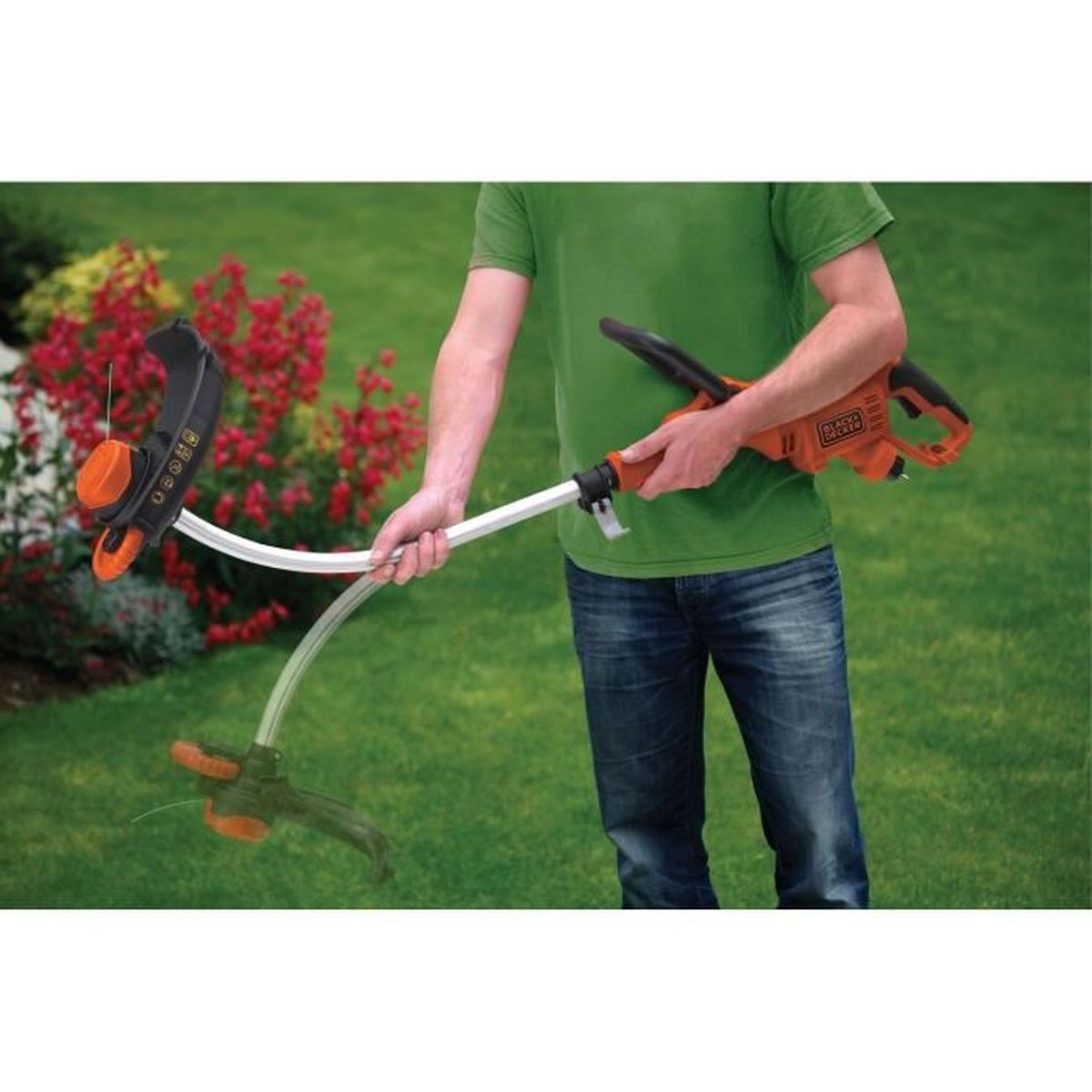 Ein Mann im grünen T-Shirt und Jeans hält einen orange-schwarzen Rasentrimmer in einem Garten mit roten Blumen.