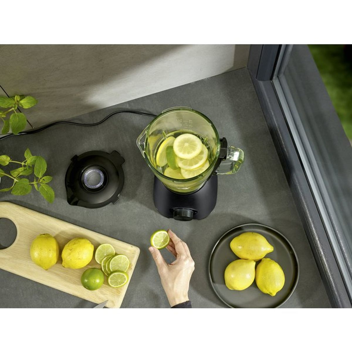 Hand hält Limettenscheibe nahe einem Mixer mit Zitronen, Schneidebrett und Tellern auf grauer Arbeitsplatte.