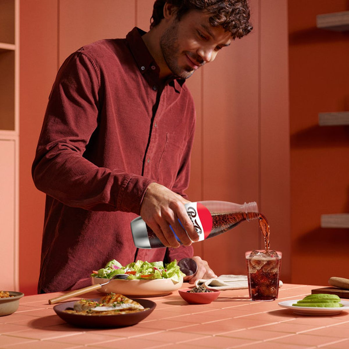 Ein Mann im roten Hemd gießt Cola aus einer Flasche in ein Glas auf einem Tisch mit Salat und Fleisch.