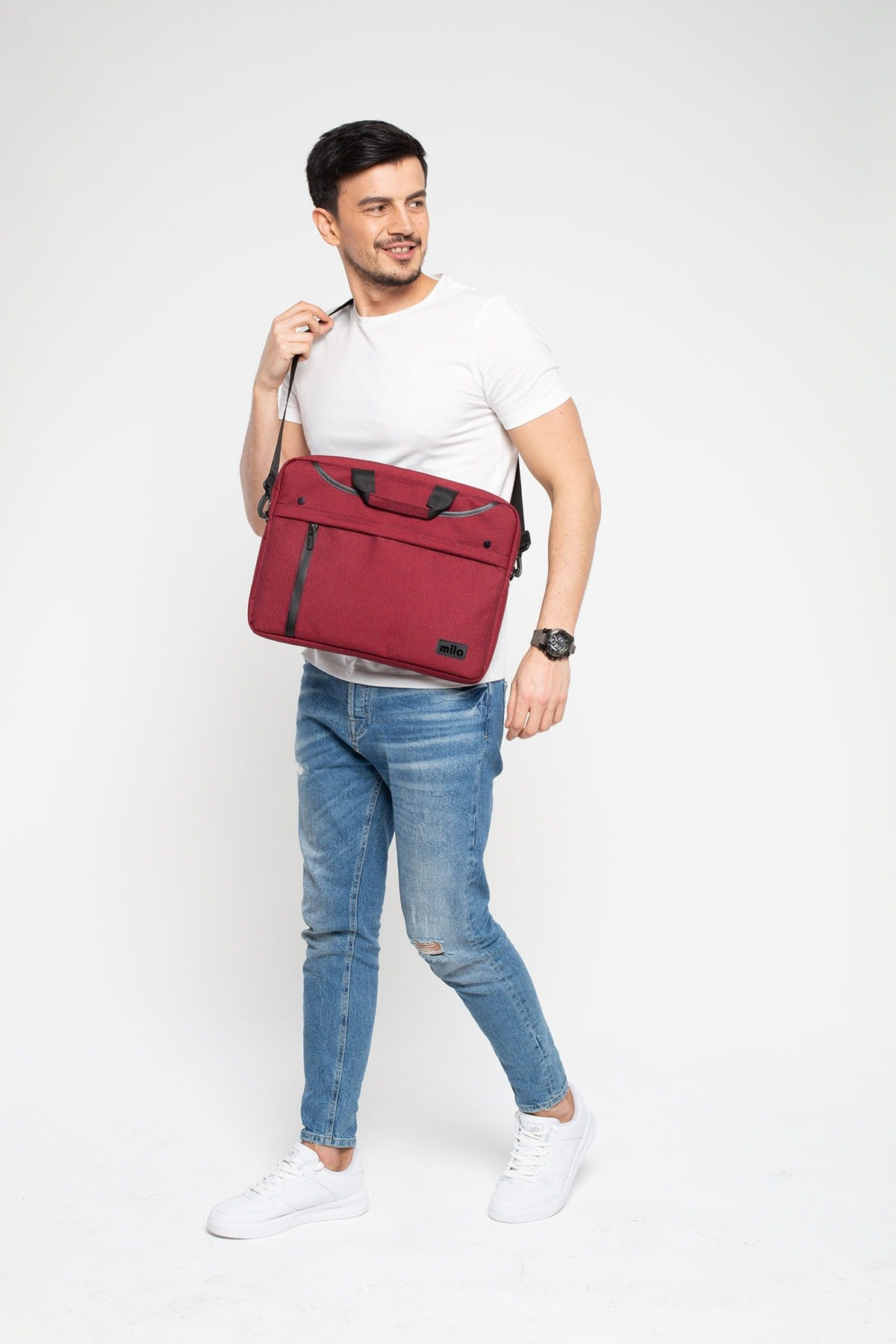 Beyaz tişörtlü ve kot pantolonlu bir adam, beyaz arka planda omzunda kırmızı bir laptop çantası taşıyor.