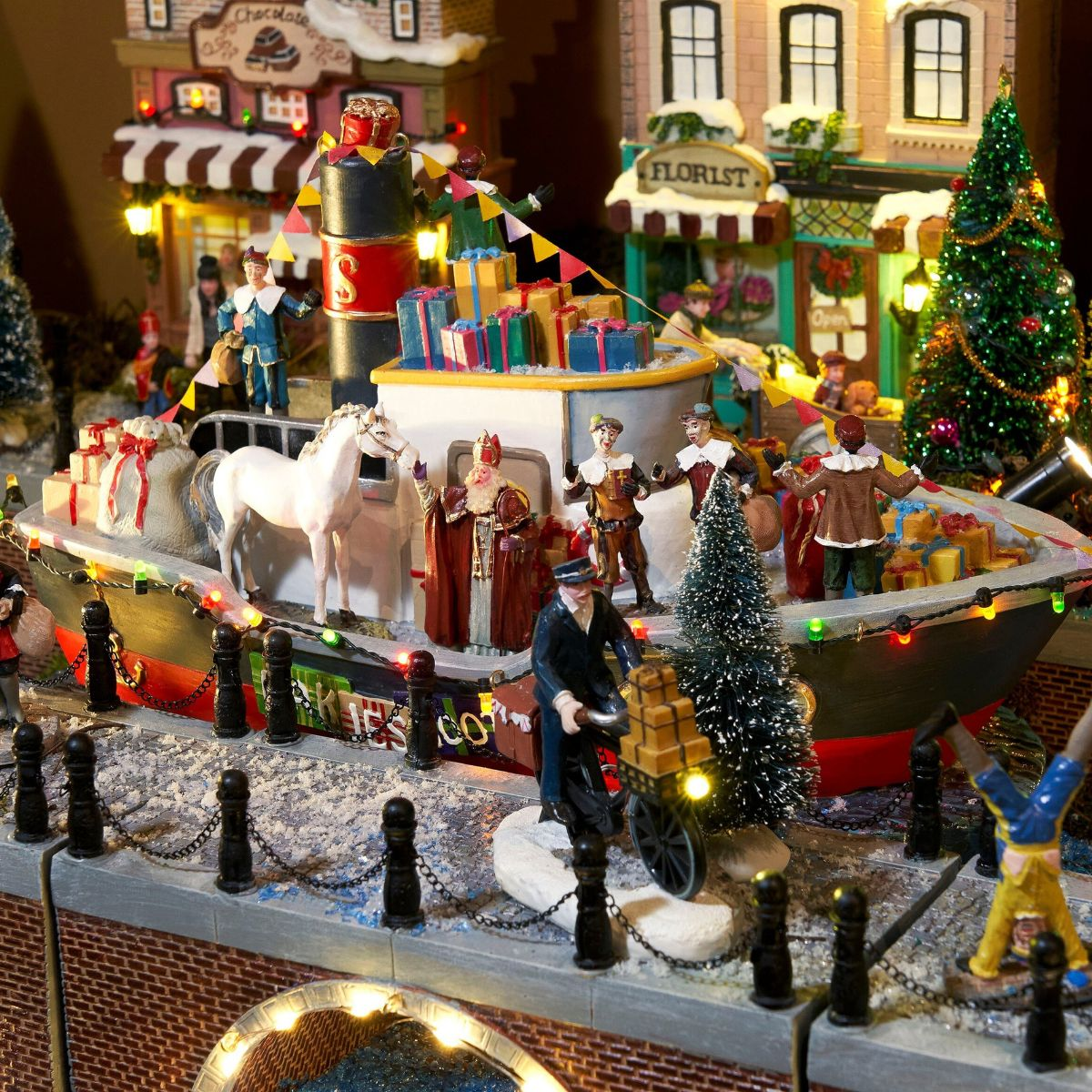 Een verlicht kerstdorpmodel toont een botenparade met figuren zoals de Kerstman, cadeautjes en kerstbomen.