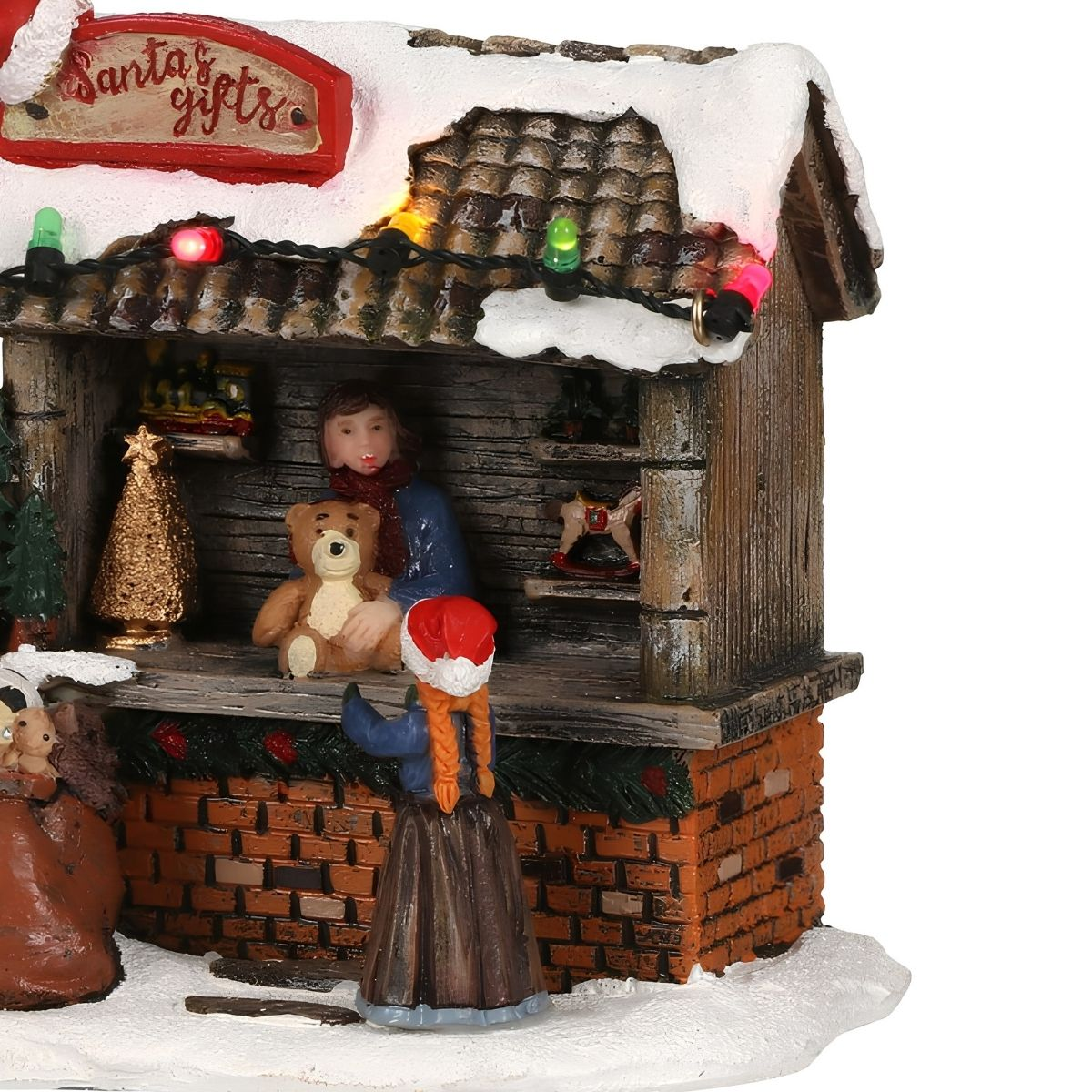 Een kerstdorpfiguur toont een winkel met een verkoper die een teddybeer vasthoudt en een kind ervoor.