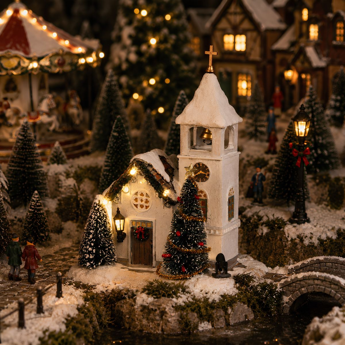 Een feestelijk, besneeuwd kerstdorp tafereel met een verlichte kerk, bomen, beeldjes en een brug.