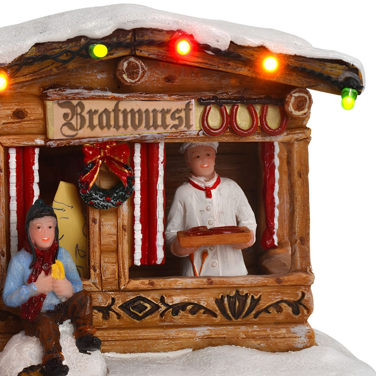 Christmas village figurine of a bratwurst stall with a chef and customer in snow with colorful lights.