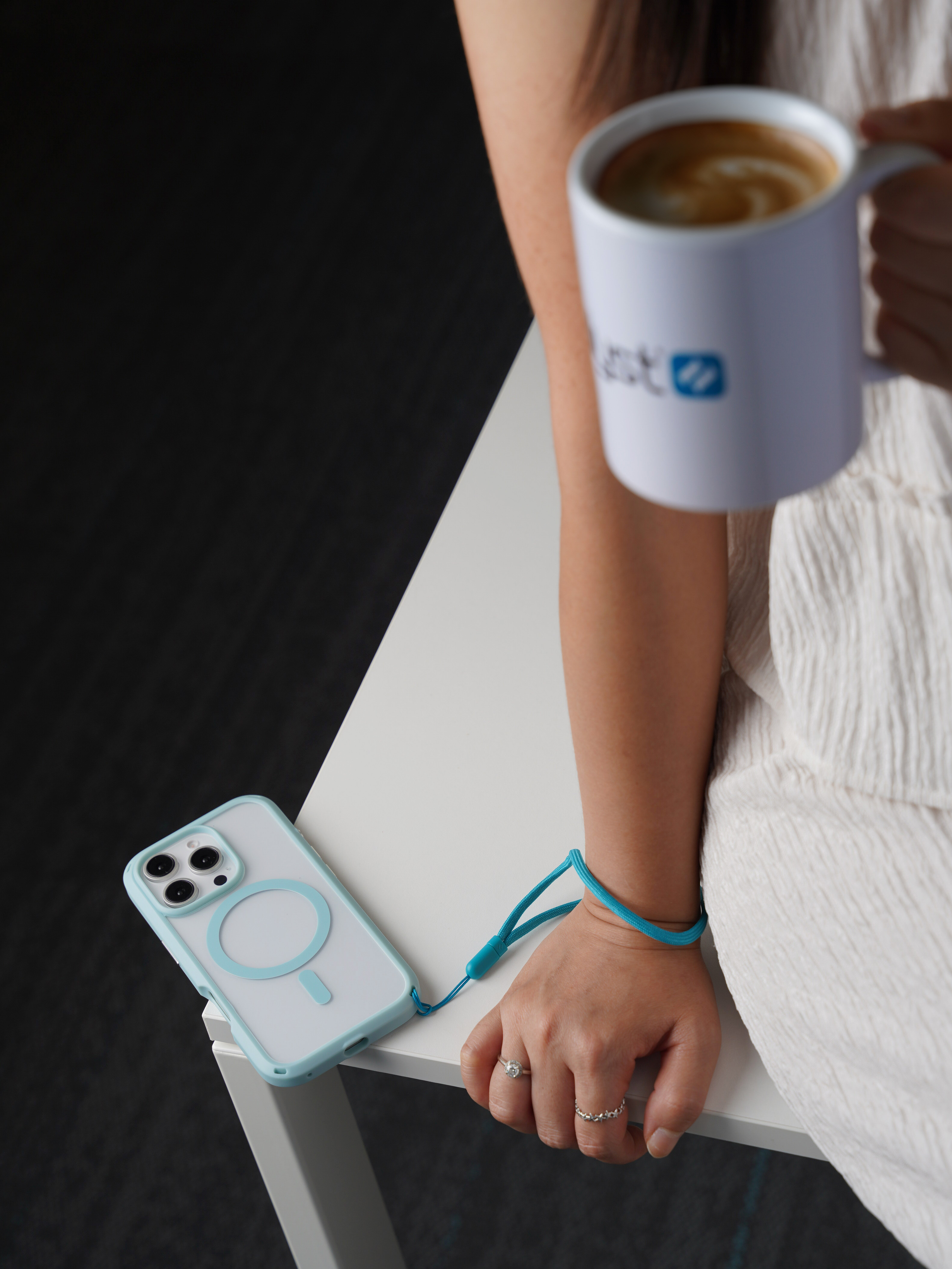 Eine Frau hält eine Kaffeetasse, während ein Smartphone mit hellblauer Hülle und Umhängeband auf dem weißen Tisch liegt.
