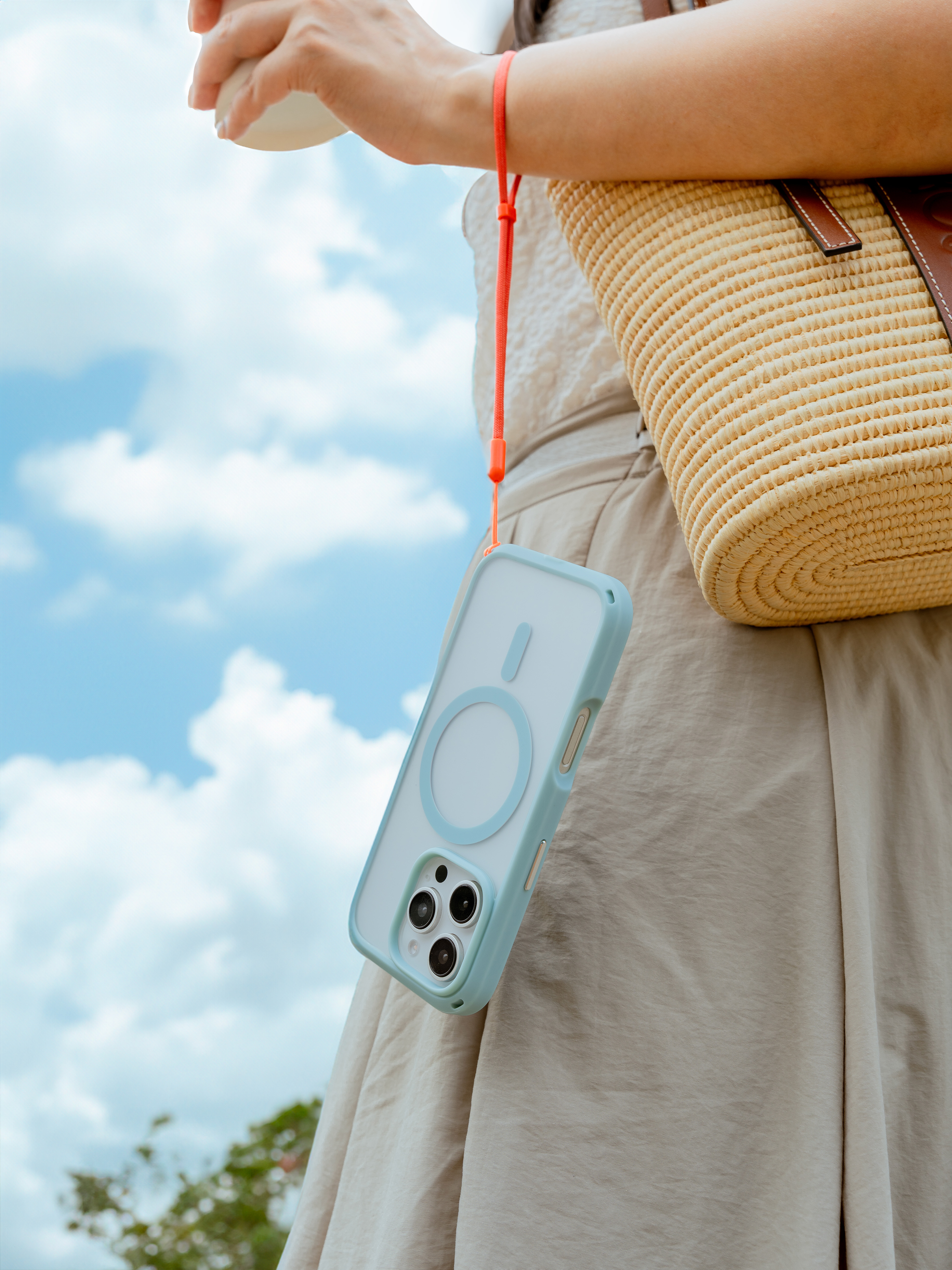 Eine Frau trägt einen Strohsack und ein blaues Smartphone mit einem orangen Umhängeband vor einem Wolkenhimmel.