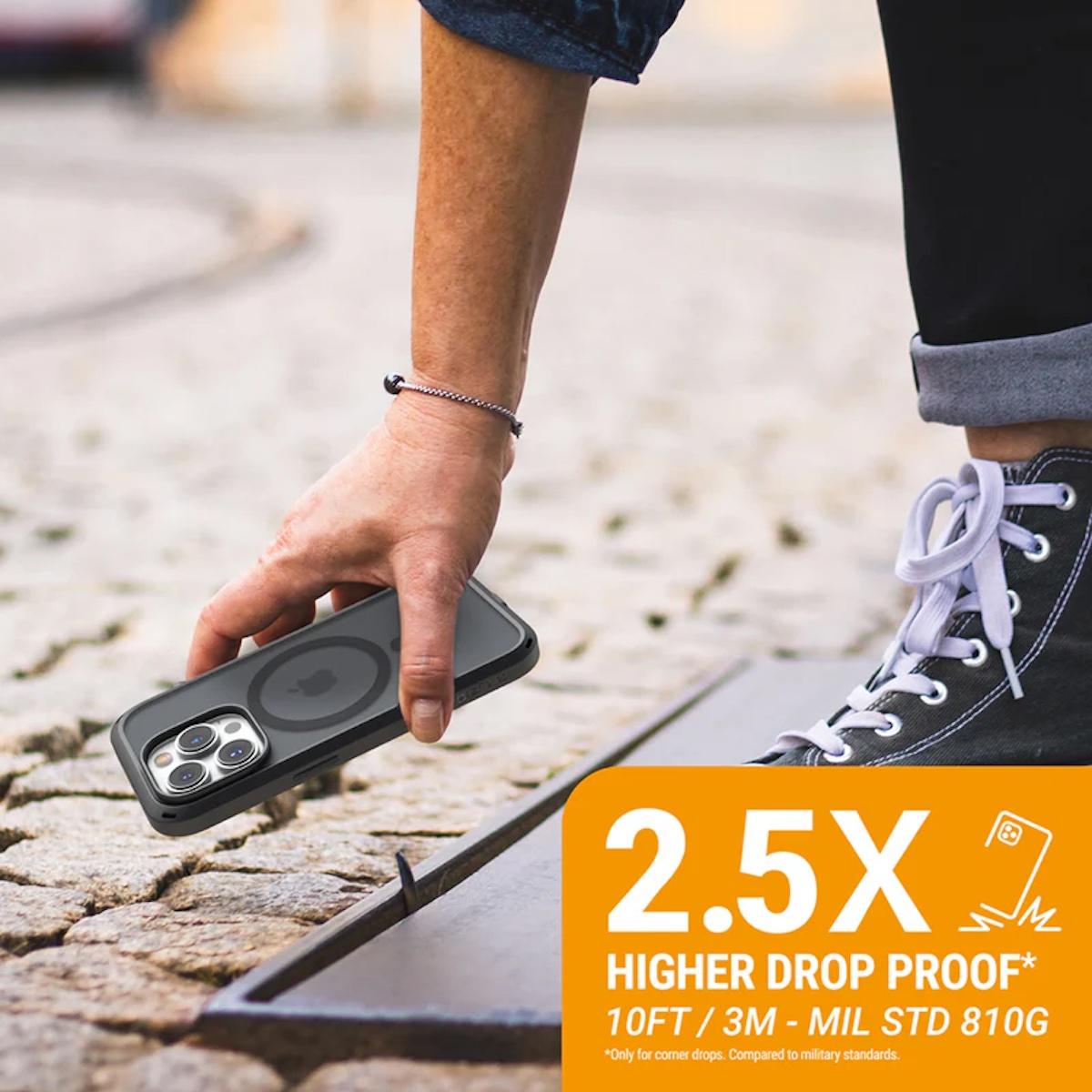 Eine Person hält ein Handy in stabiler Hülle über einem Steinweg, mit orangem Overlay für 2,5x Sturzschutz.