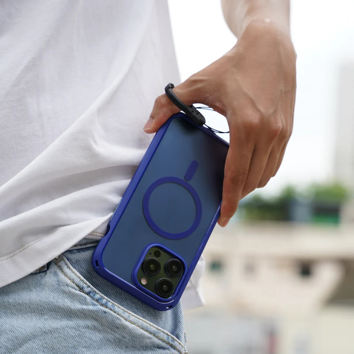 Eine Person trägt ein blaues Smartphone mit schwarzem Bumper-Case in der Hand, nah an Jeans und weißem Shirt.