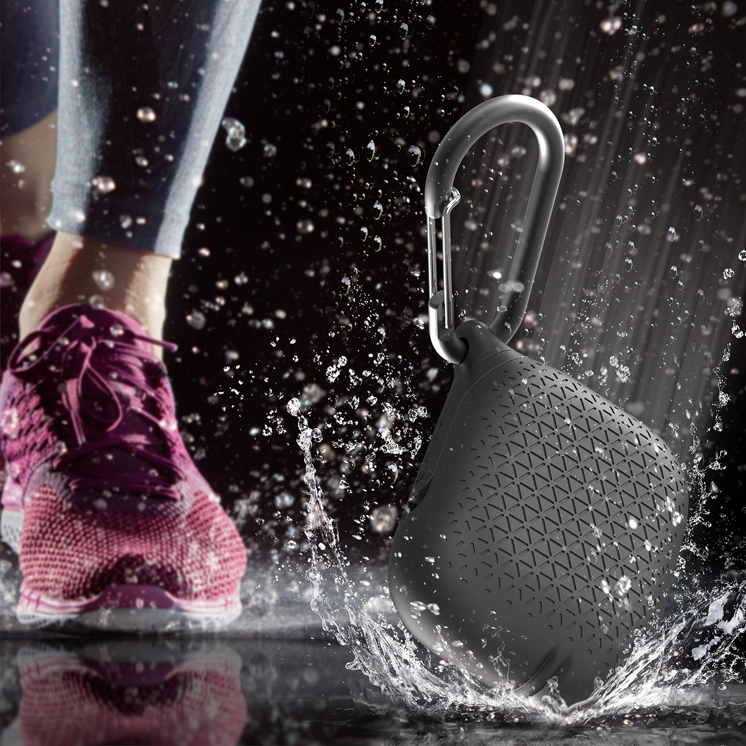 Ein schwarzer Lautsprecher mit Karabiner im Wasserspritzer, im Hintergrund ein pinker Laufschuh.