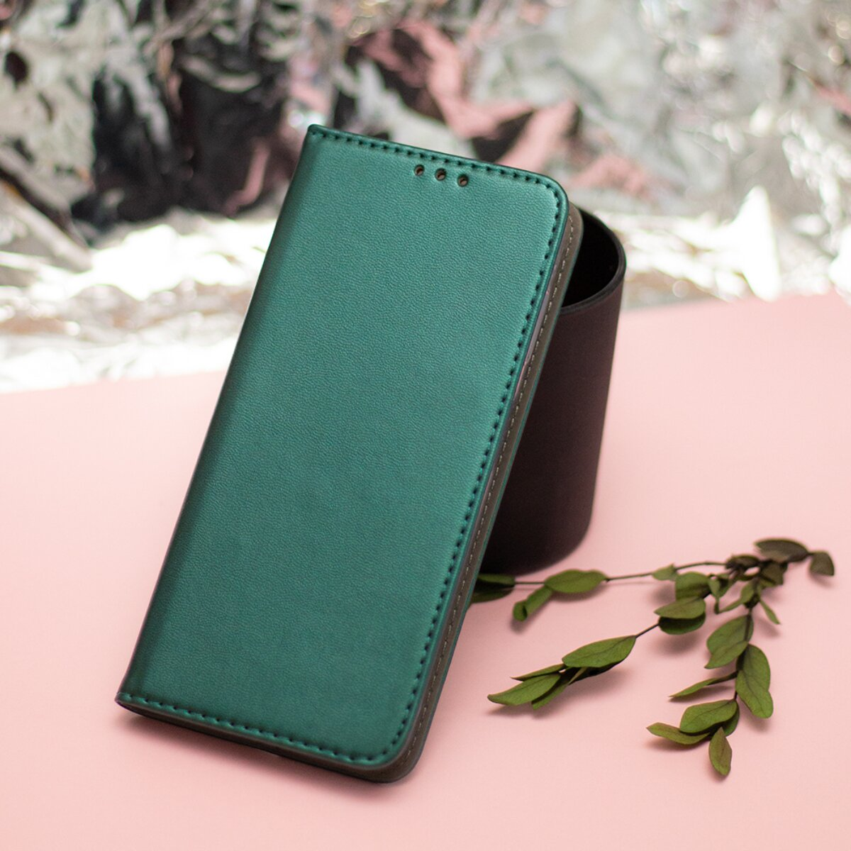 Eine metallic-grüne Leder-Handyhülle steht aufrecht auf einer rosa Oberfläche mit einem kleinen blättrigen Zweig.