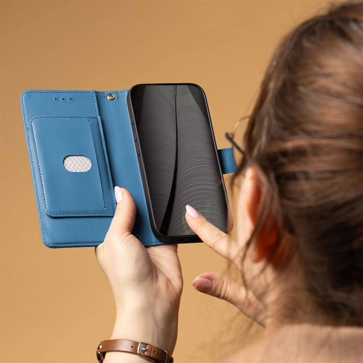 Eine Person hält eine blaue Leder-Handyhülle und tippt auf den Bildschirm vor einfarbigem Hintergrund.