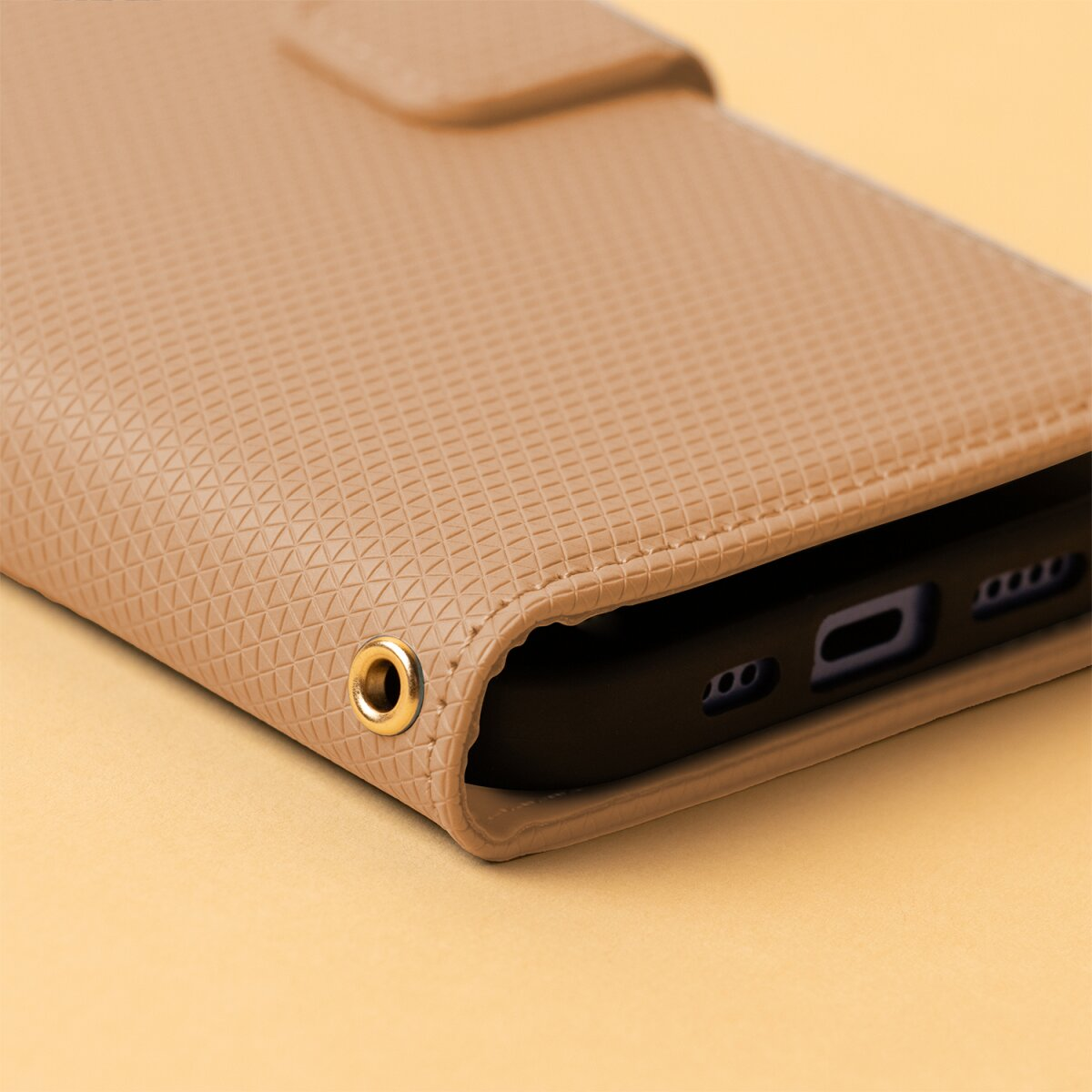 Ein beiges, strukturiertes Handy-Etui aus Leder mit einem kleinen goldenen Ring auf einer beigen Oberfläche.
