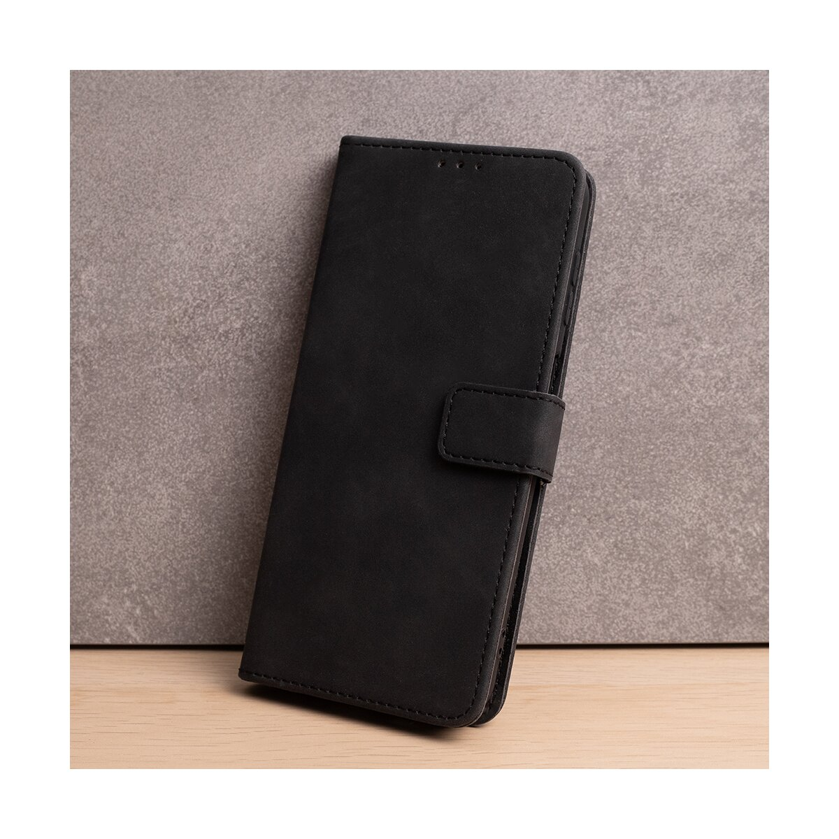 Eine schwarze Handytasche im Buchformat steht auf einer Holzoberfläche vor einer grauen Wand.