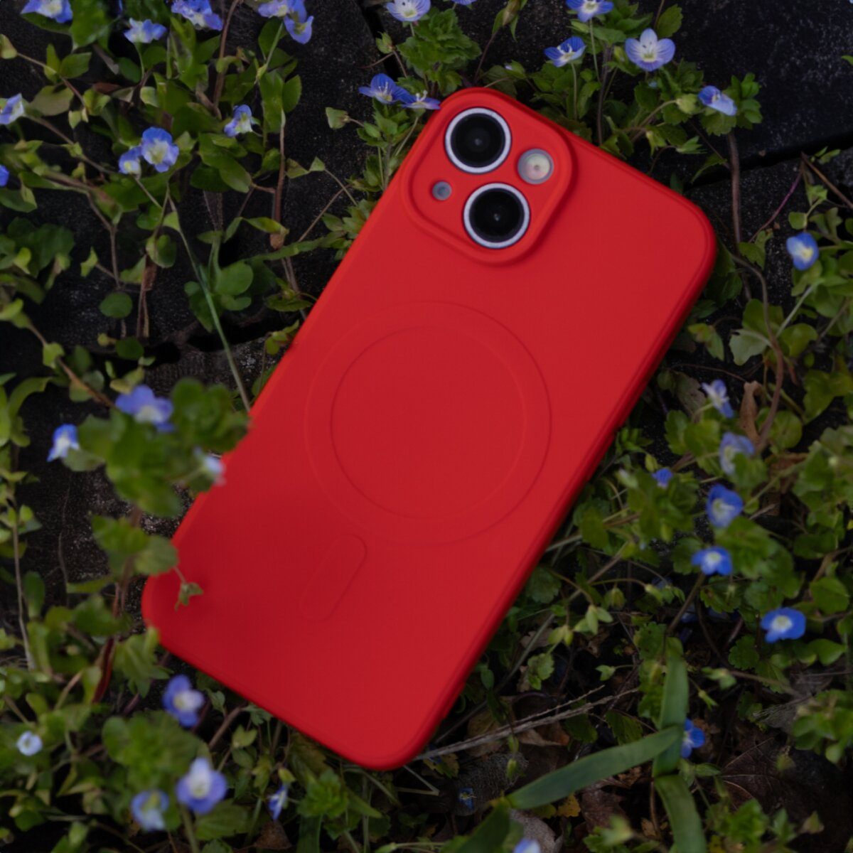 Ein rotes Smartphone mit Hülle liegt im Gras zwischen kleinen blauen Blumen.