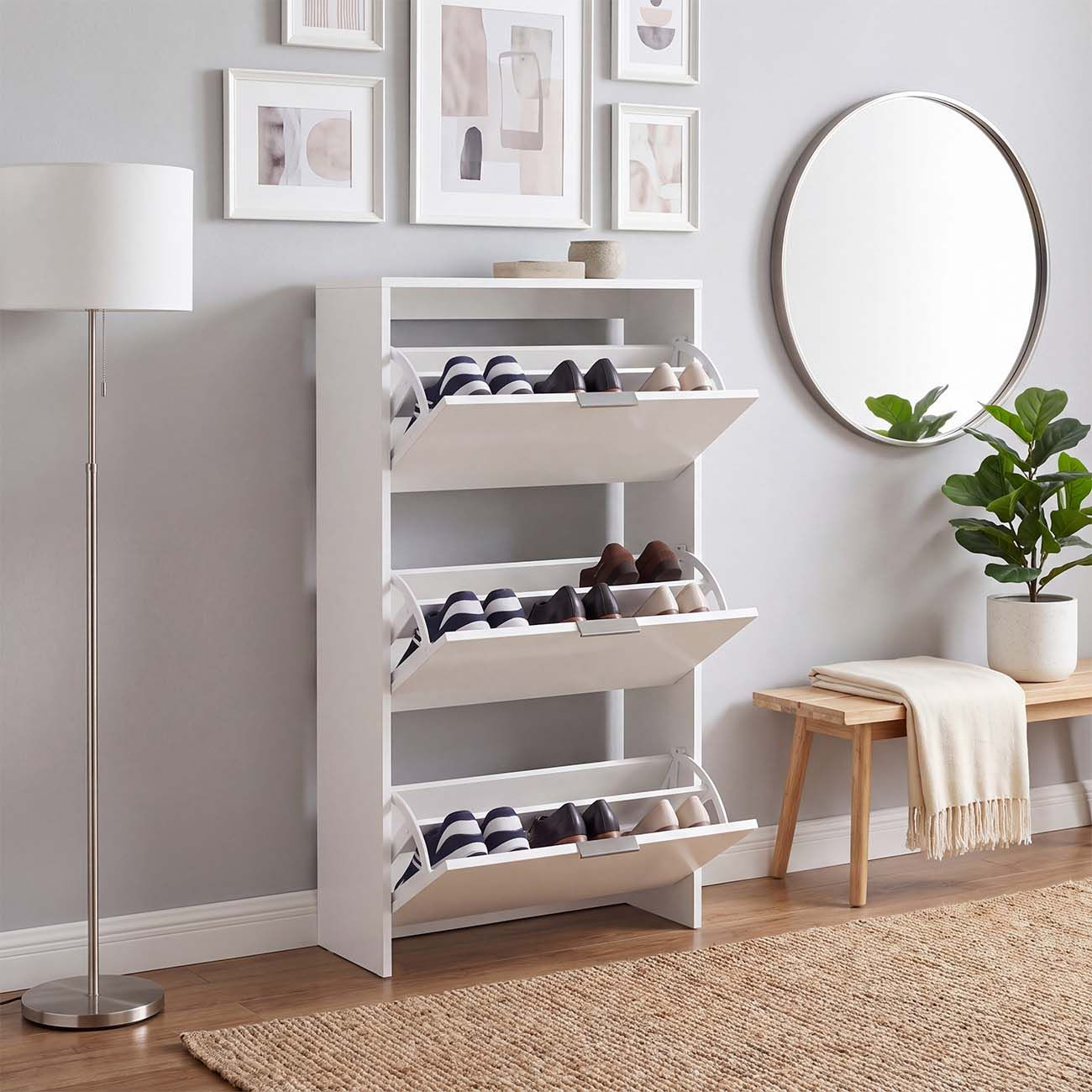 Un zapatero blanco con tres cajones abatibles llenos de zapatos en un pasillo, rodeado de decoraciones y una lámpara.