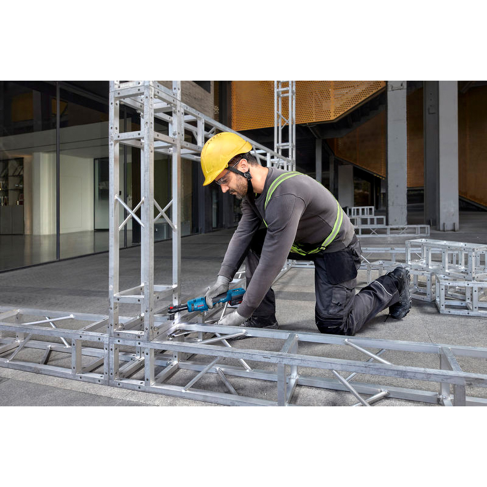 Bauarbeiter mit gelbem Helm und Schutzbrille kniet auf einer Baustelle und bedient ein Elektrowerkzeug an Metallrahmen.