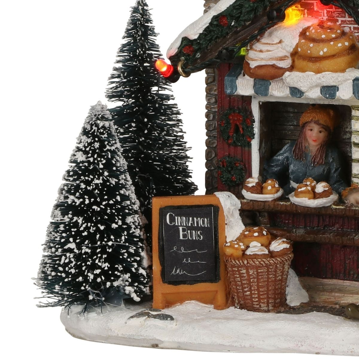 Een besneeuwde kerstkraam met kaneelbroodjes, bomen, een bord en een verkoper.