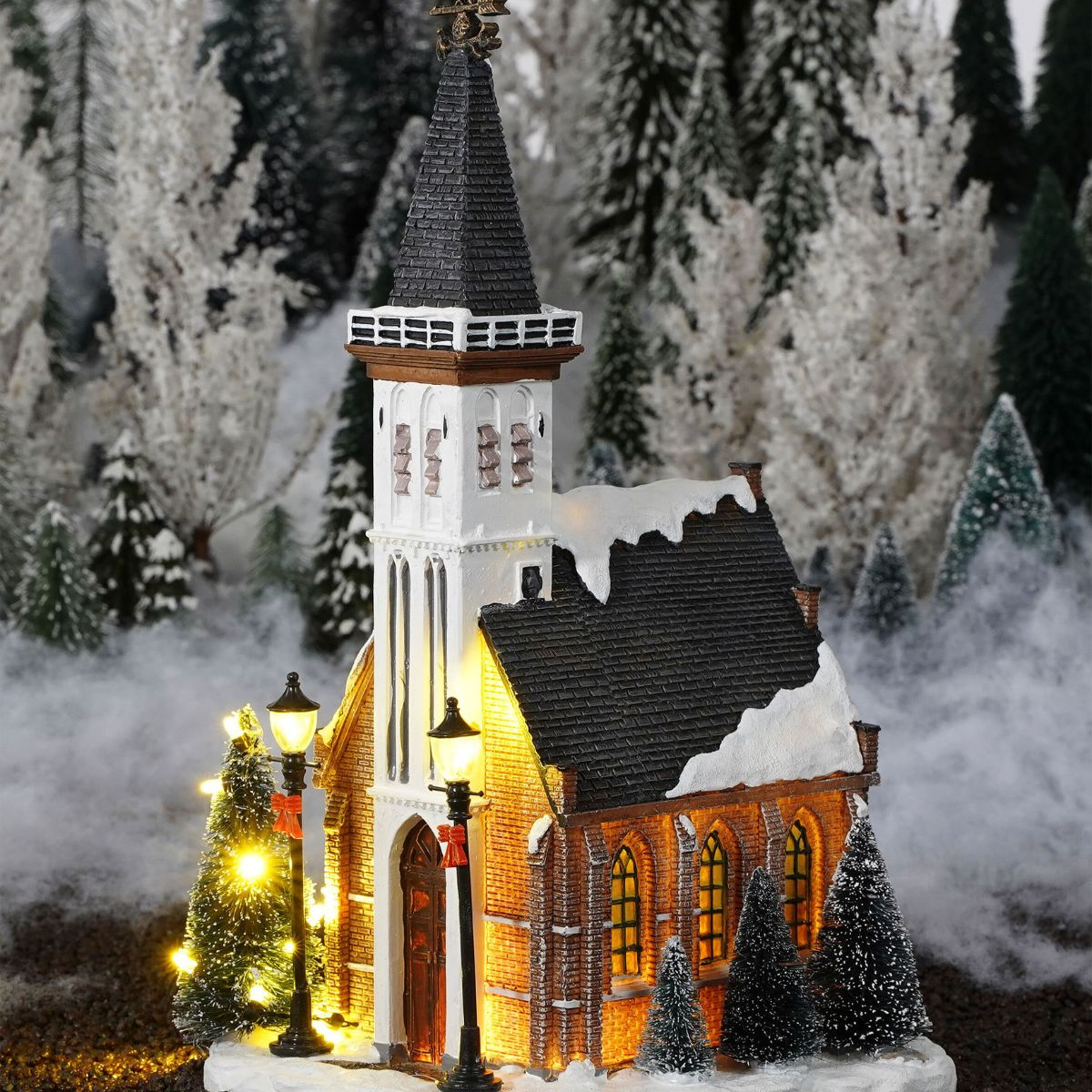 A glowing miniature church with snow-capped roofs stands among lit Christmas trees and a snowy forest backdrop.