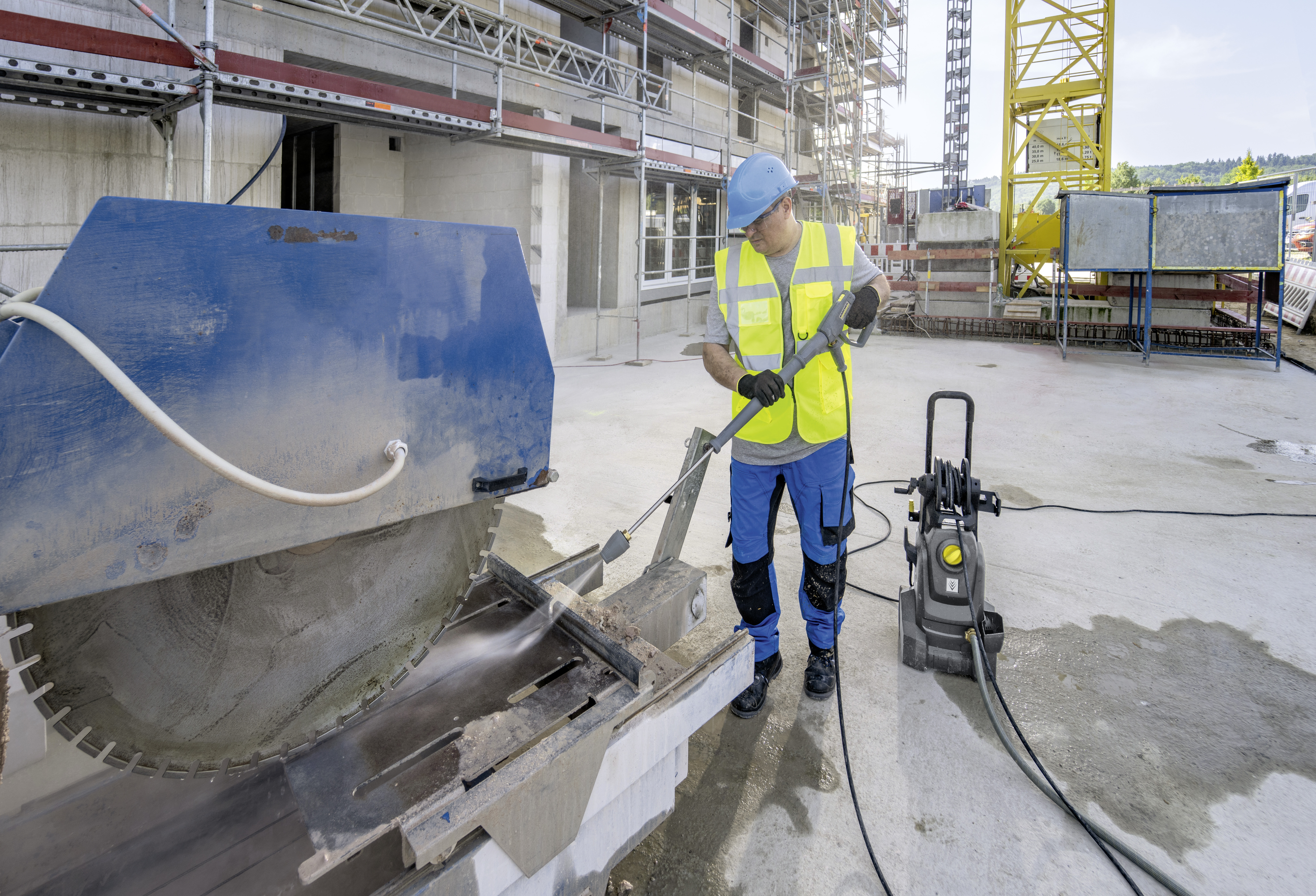 Bauarbeiter reinigt eine große Steinsäge mit einem Hochdruckreiniger auf einer Baustelle.