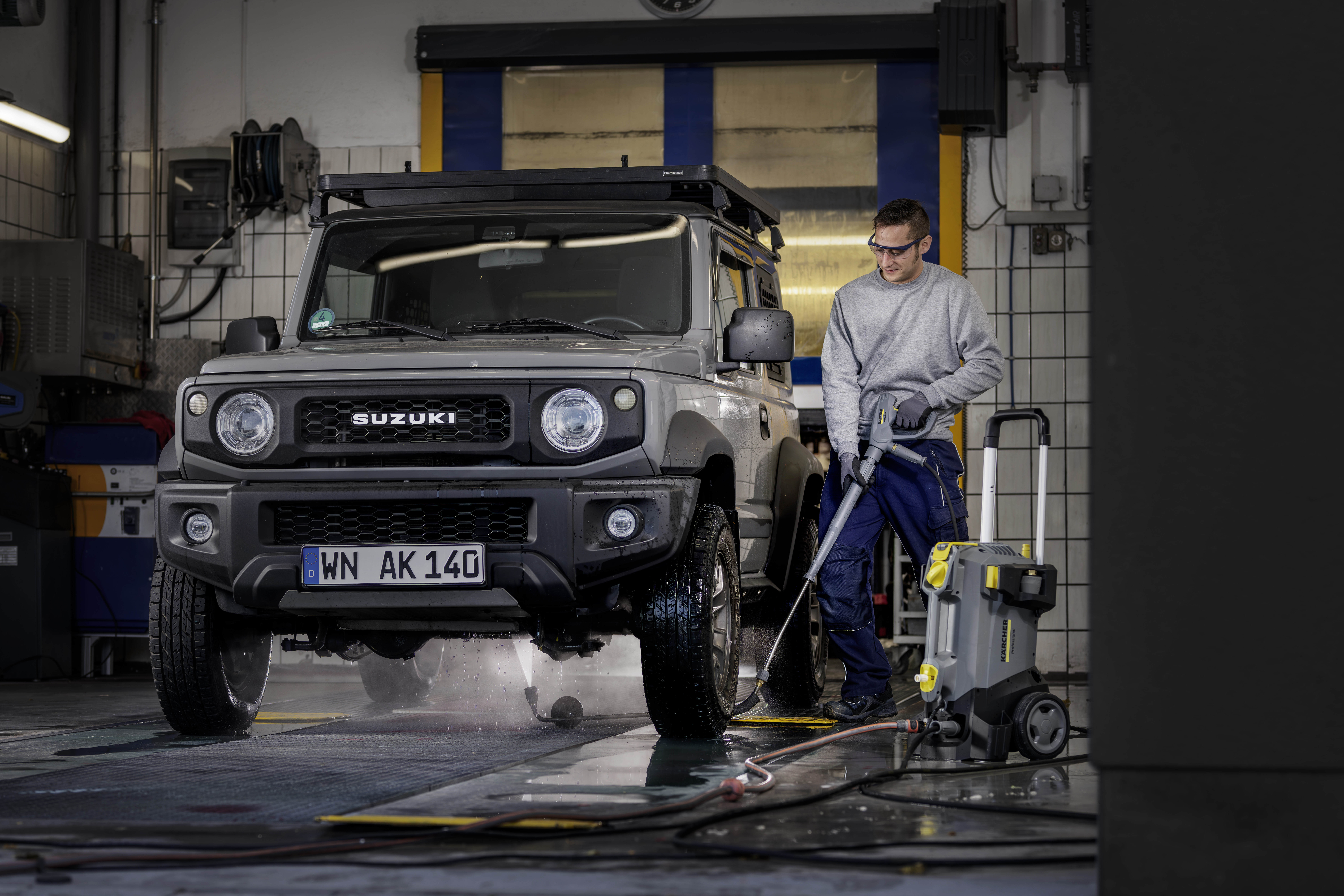 Ein Mann wäscht einen grauen Suzuki Geländewagen mit einem Hochdruckreiniger in einer Werkstatt.