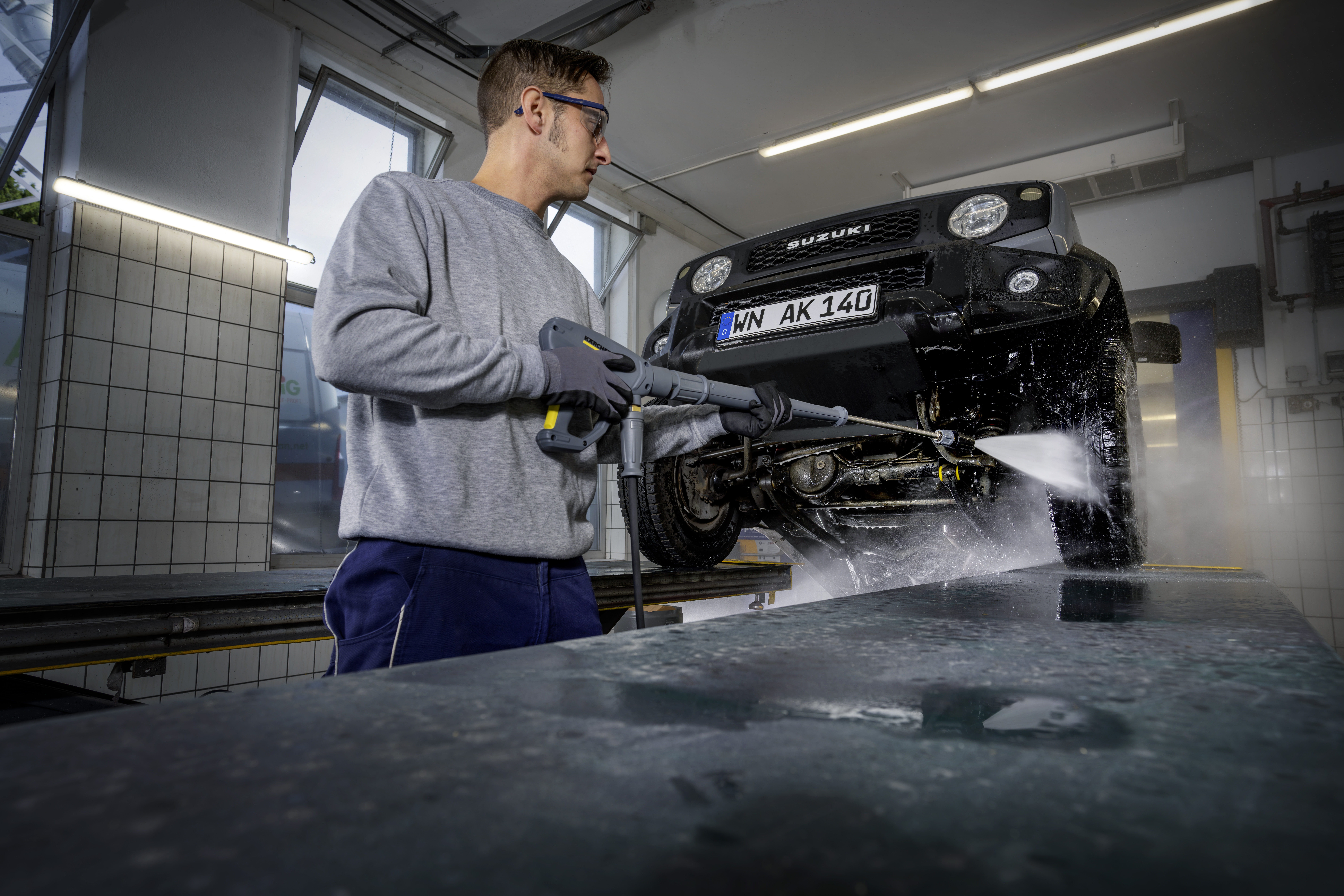 Ein Arbeiter reinigt mit einem Hochdruckreiniger das Unterteil eines schwarzen Suzukis in einer Werkstatt.