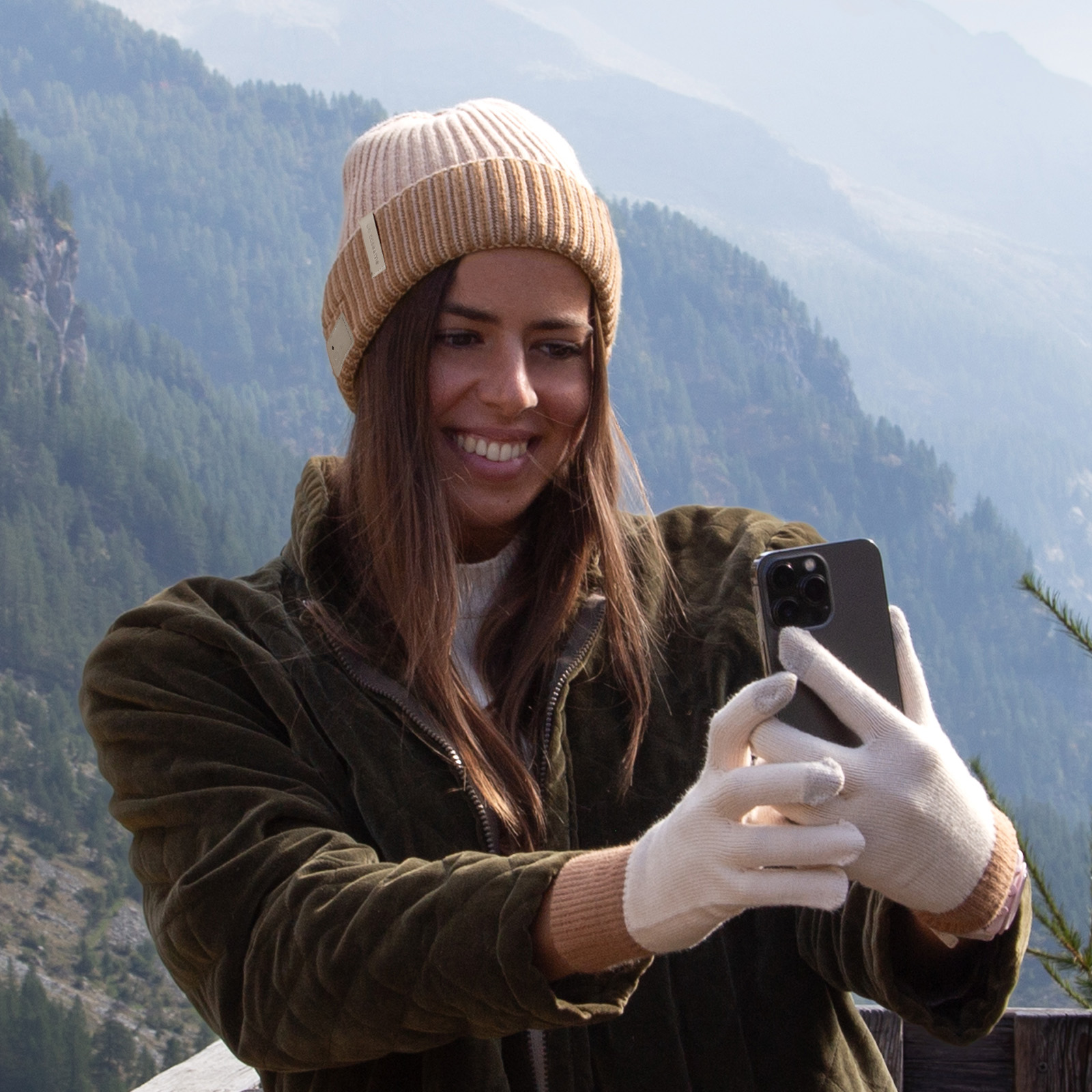 Una mujer sonriente con gorro y chaqueta se hace un selfie frente a un fondo de bosque montañoso.