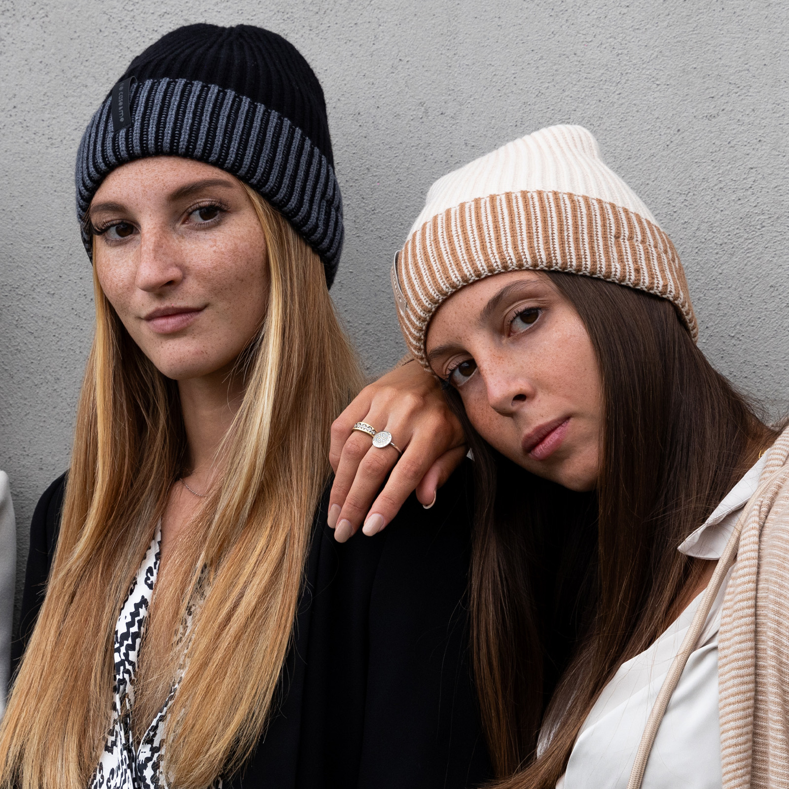 Dos mujeres con cabello largo y gorros de punto posan frente a una pared gris.