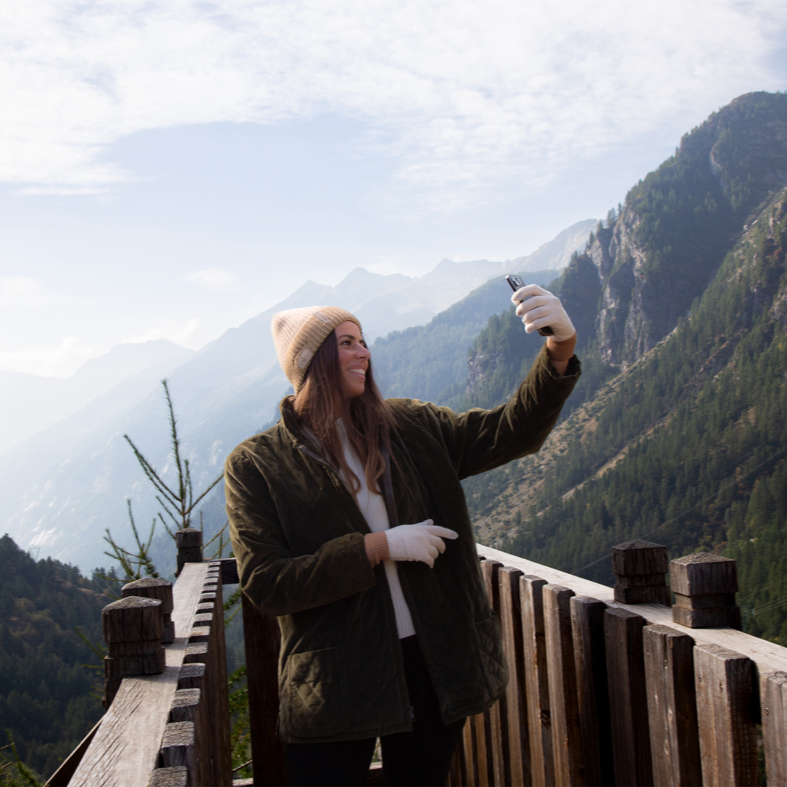 Una mujer con gorro de invierno toma un selfi en un balcón de madera con vista a la montaña y al bosque.