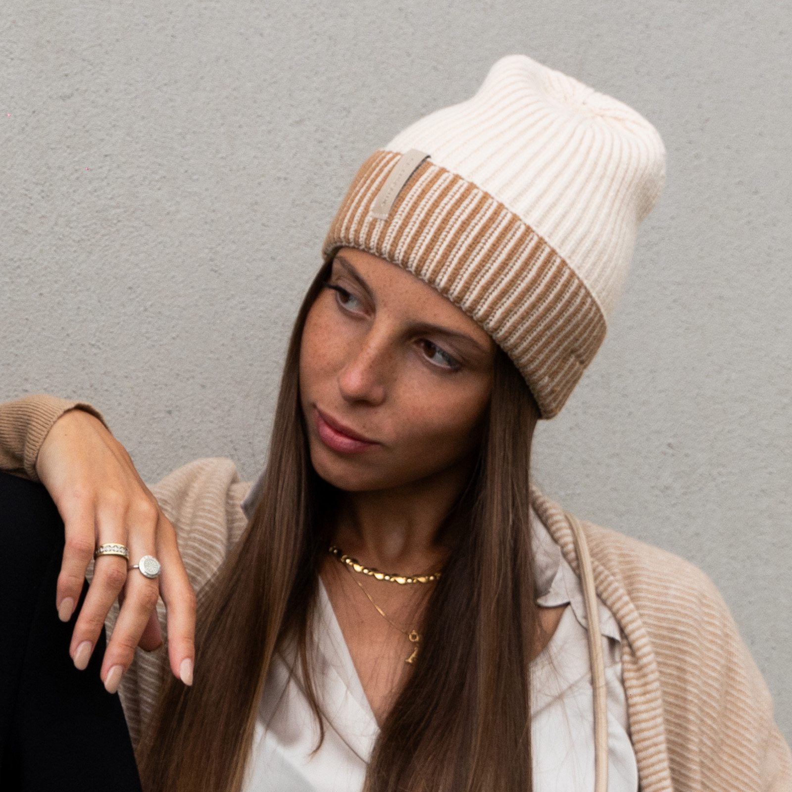 Una mujer con cabello castaño largo que lleva un gorro crema y marrón, cárdigan y joyas contra una pared gris.