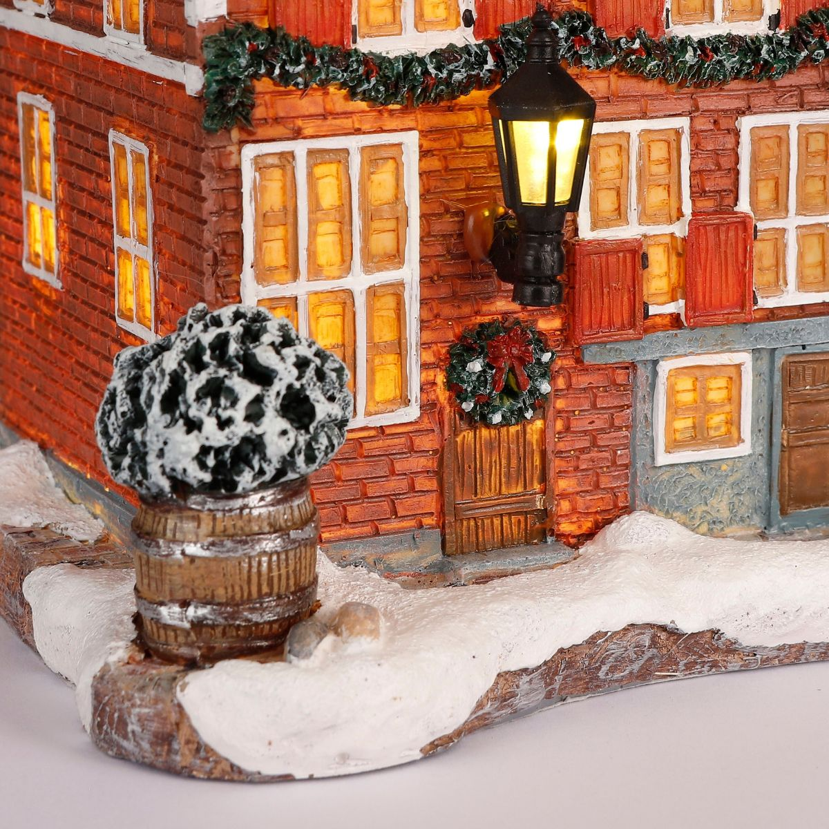 Verlicht kerststenen huis met sneeuw, lantaarn, krans en houten vat op de voorgrond.