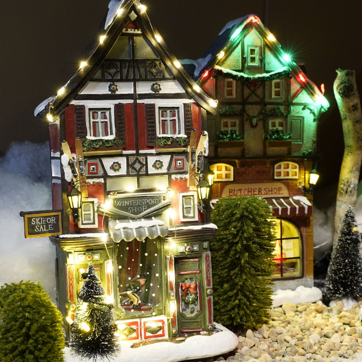 Un village d'hiver miniature illuminé avec un magasin de ski, enneigé, entouré de petits arbres et de cailloux.