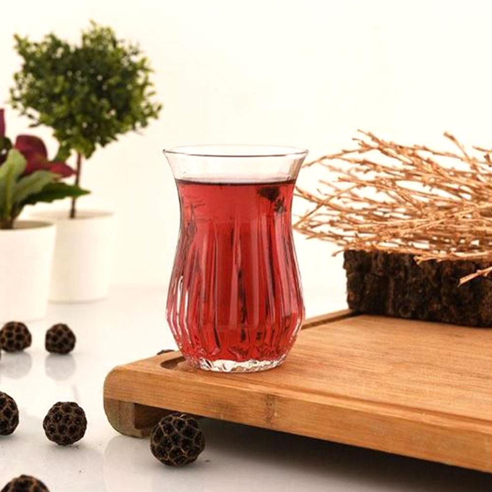 Kırmızı içecekle dolu bir bardak ahşap tahta üzerinde duruyor, küçük koyu küpler ve süslemelerle çevrili.