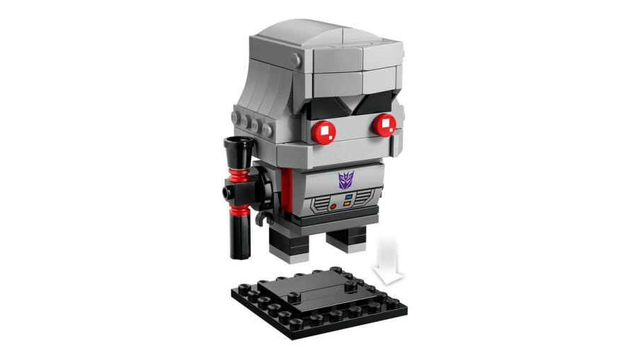 Beyaz arka planda siyah tabanın üzerinde kırmızı silahlı gri ve siyah Lego Megatron figürü.