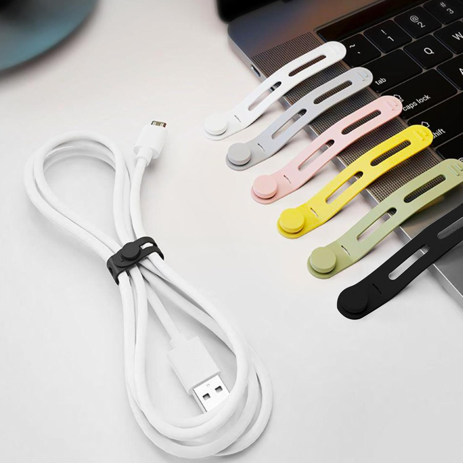 Ein weißes USB-Kabel mit schwarzem Clip, neben bunten Kabelorganisatoren und einem Laptop auf einem weißen Tisch.