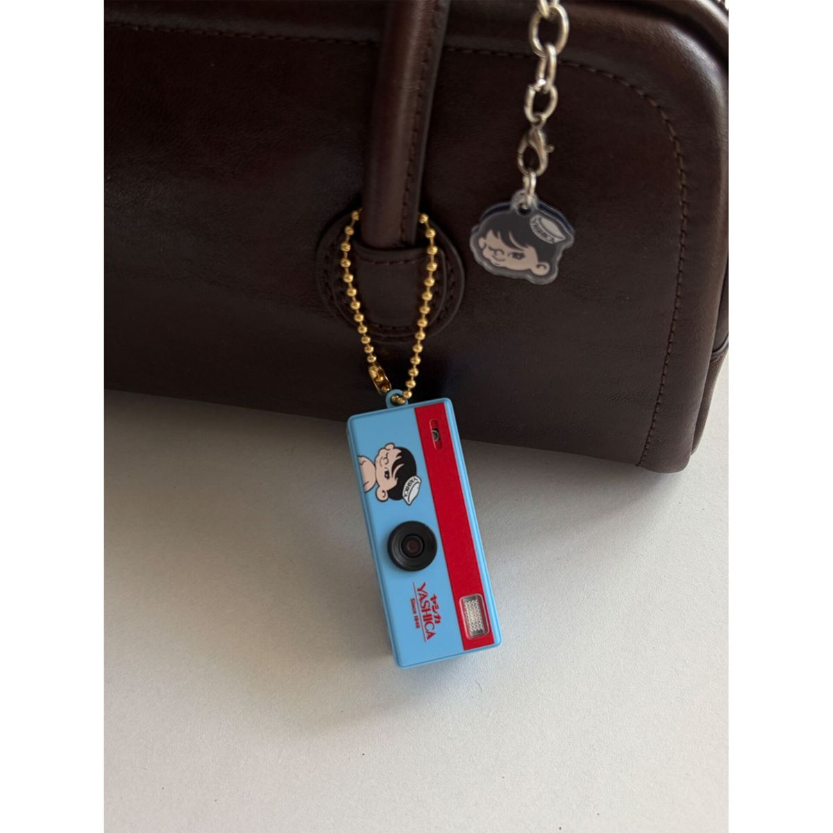 Eine blaue Yashica-Kameratasche mit Comic-Figur hängt an einer braunen Handtasche.