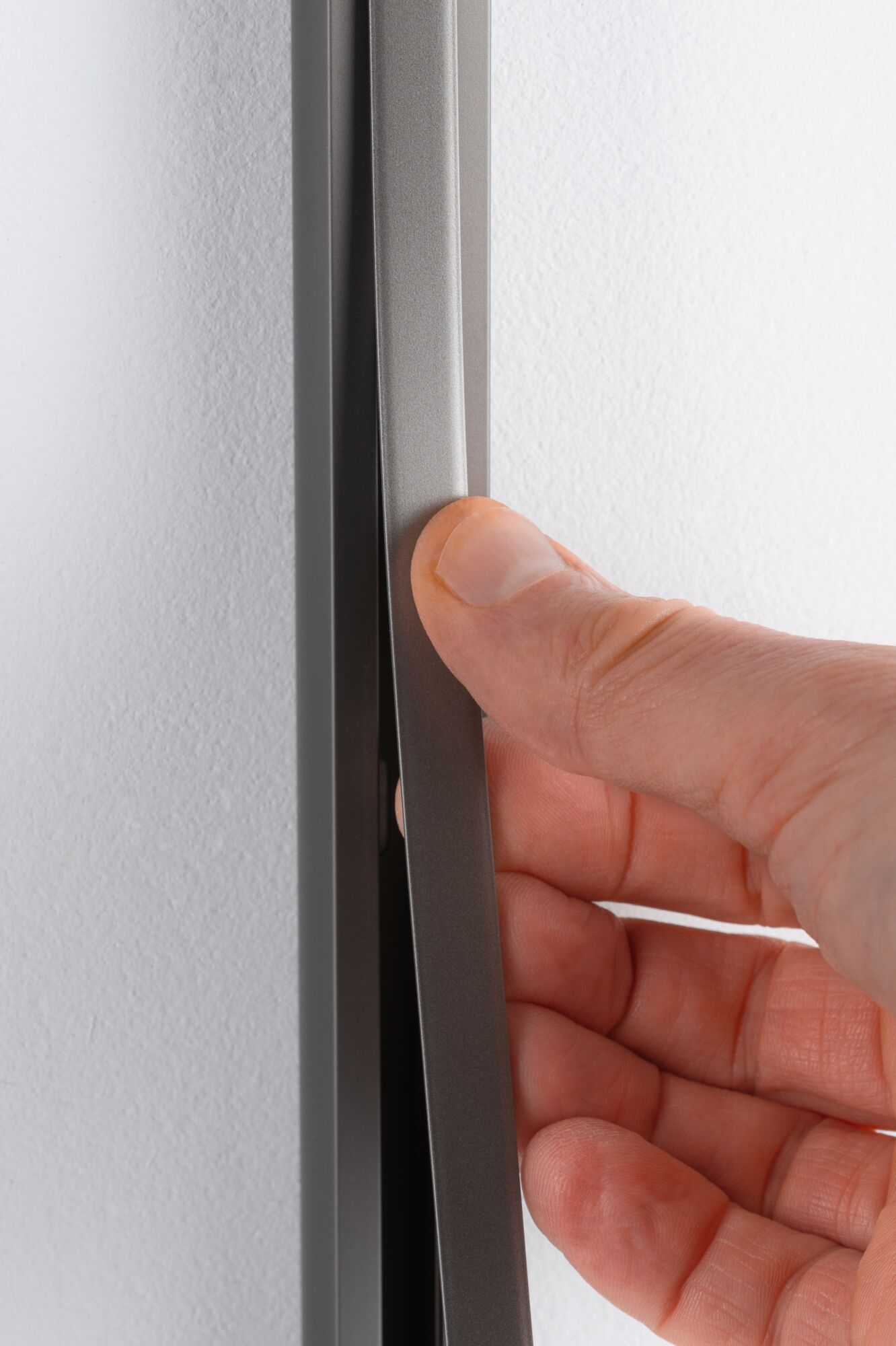 Eine Hand zieht eine graue vertikale Leiste vor einer weißen Wand weg.