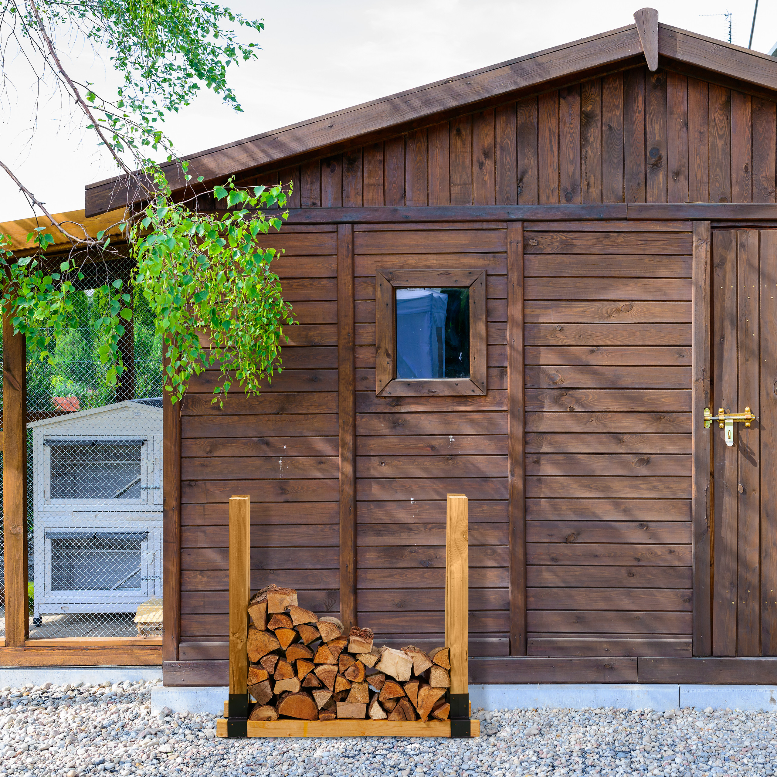 Ein Holzschuppen mit kleinem Fenster, verschlossener Tür und Holzstapelregal auf Schotter.