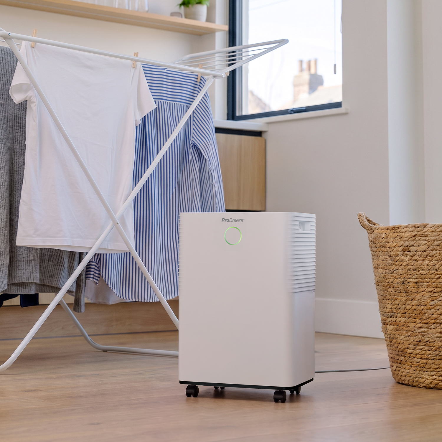 Ein weißer Luftentfeuchter steht auf einem Holzfußboden neben einem Wäscheständer und einem Korb.