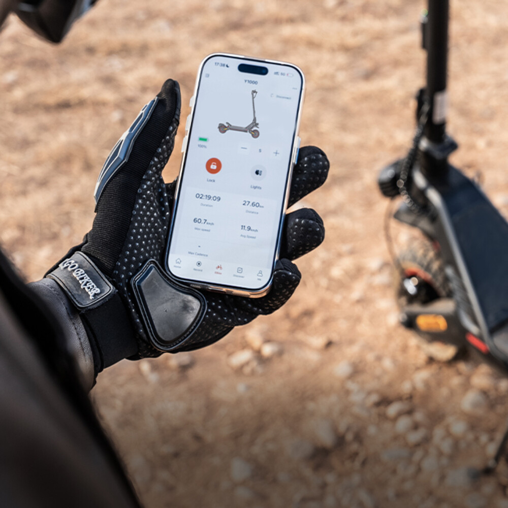 Mano con guante negro sostiene smartphone con app de patinete eléctrico, con un patinete de fondo en suelo polvoriento.