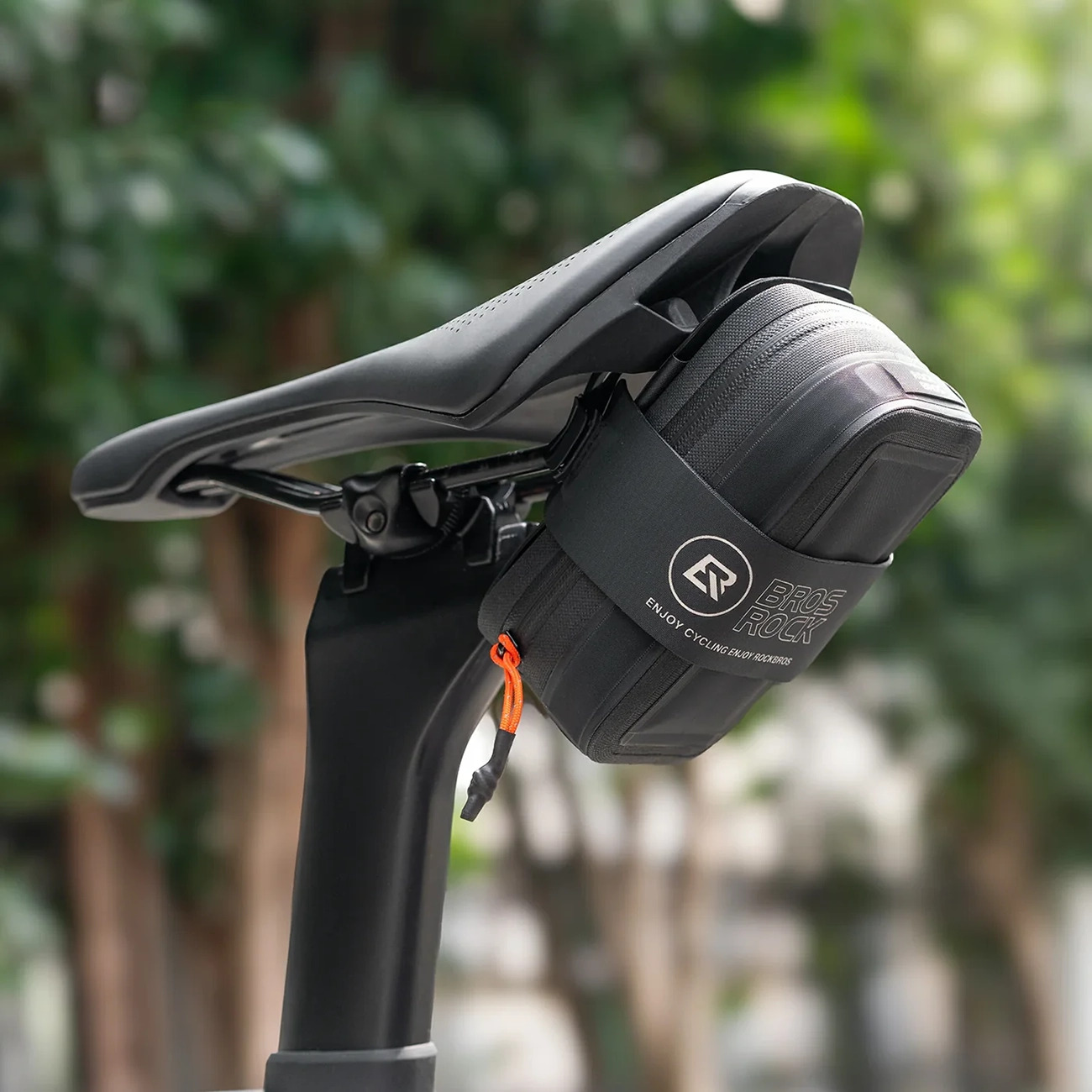 Una borsa da sella Rockbros nera è montata sotto il sedile della bicicletta, con alberi verdi sullo sfondo.