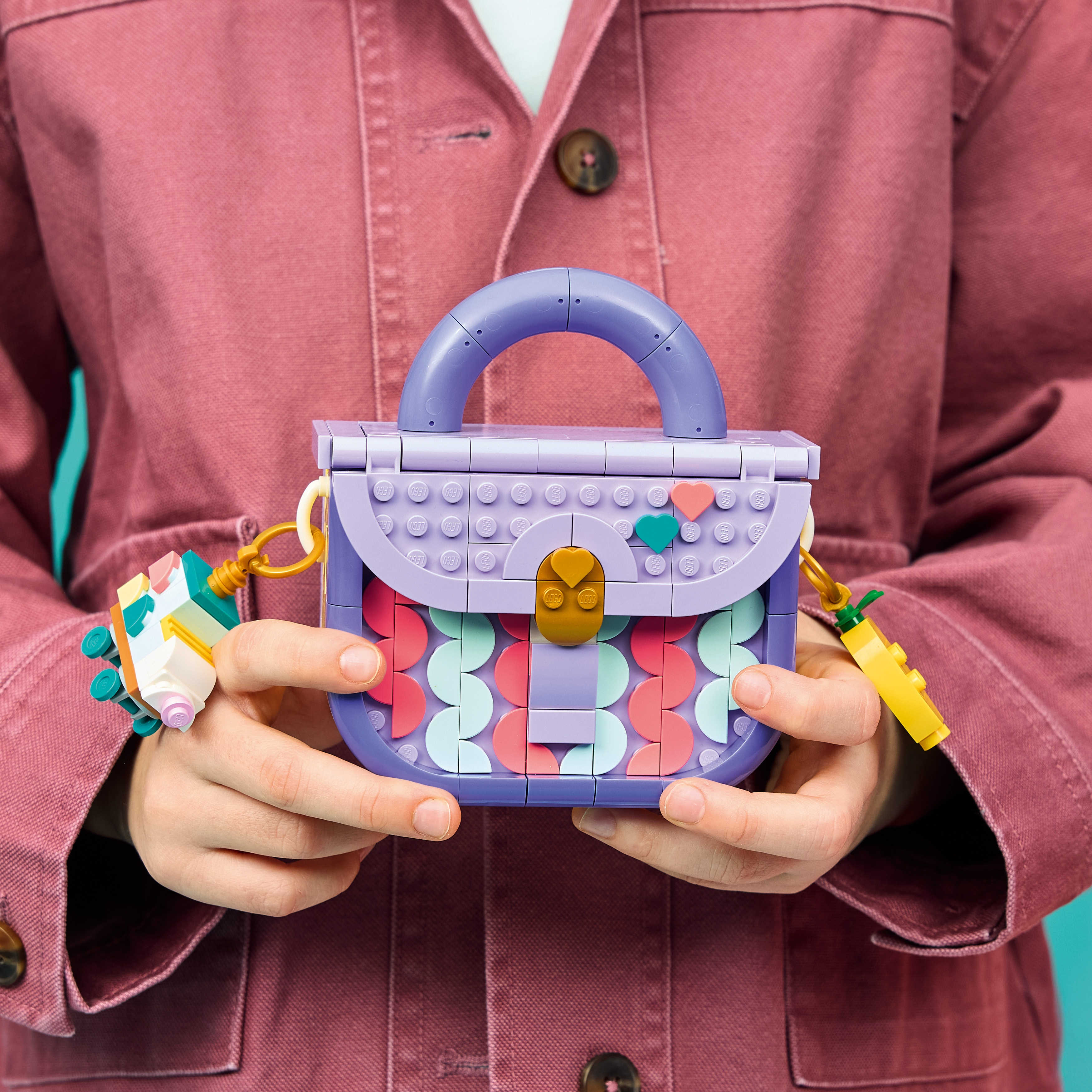 Una persona con una chaqueta rosa sostiene un bolso de juguete Lego morado con patrones coloridos.