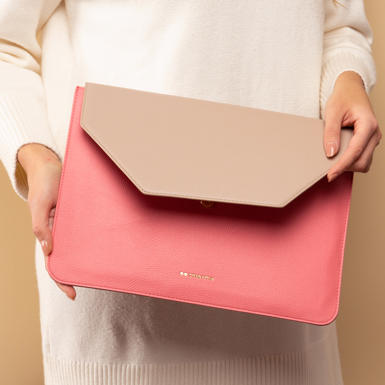 Una persona con suéter blanco sostiene una funda rosa y beige para computadora portátil con letras doradas.