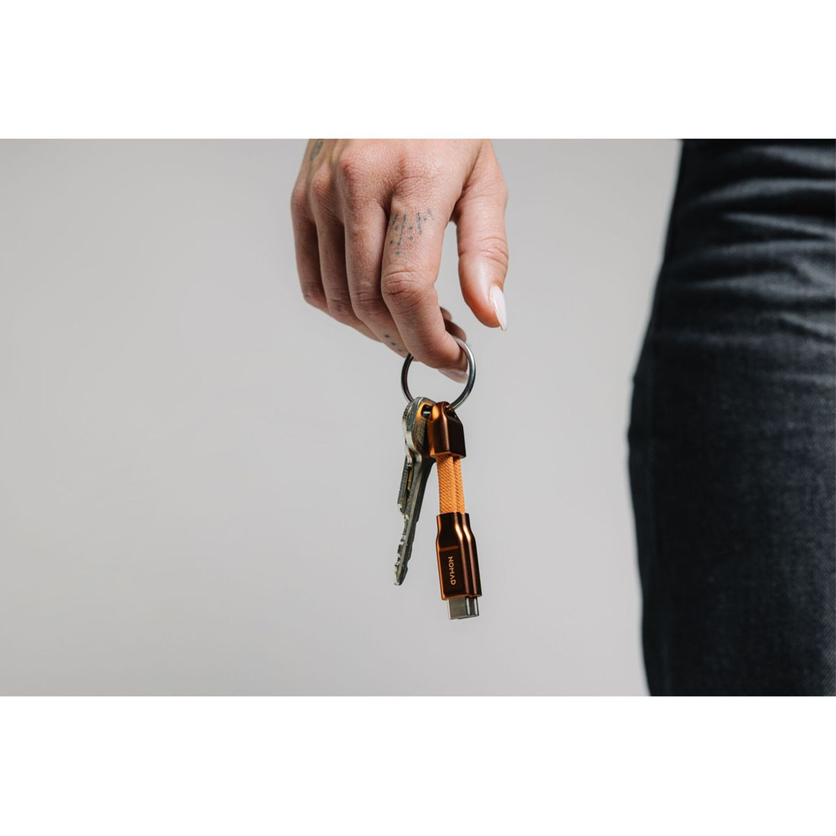 Eine Person hält einen Schlüsselring mit Schlüsseln und einem orangefarbenen USB-Kabel vor neutralem Hintergrund.