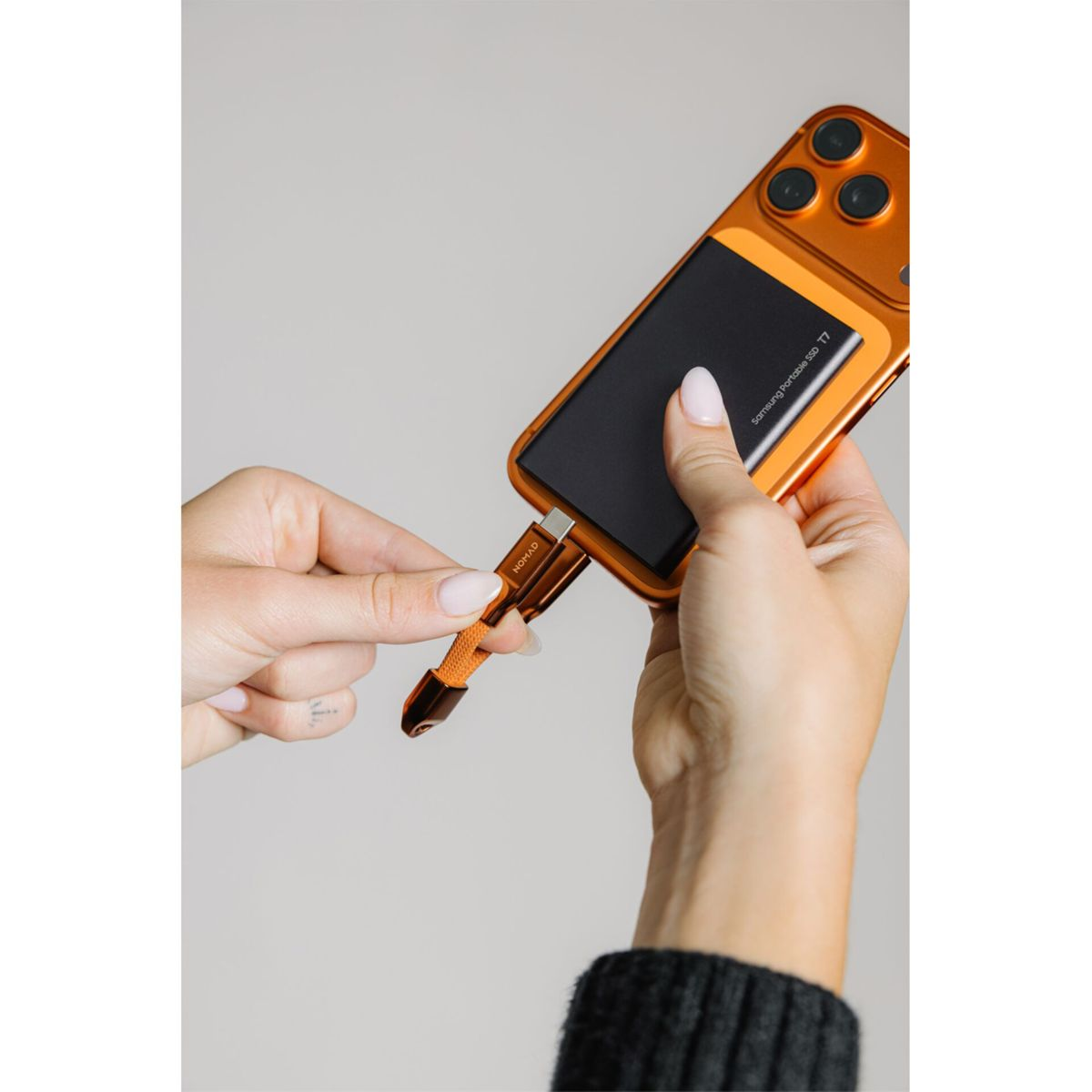 Eine Hand hält ein oranges Smartphone mit befestigter schwarzer SSD, während die andere Hand ein oranges Kabel einstöpselt.